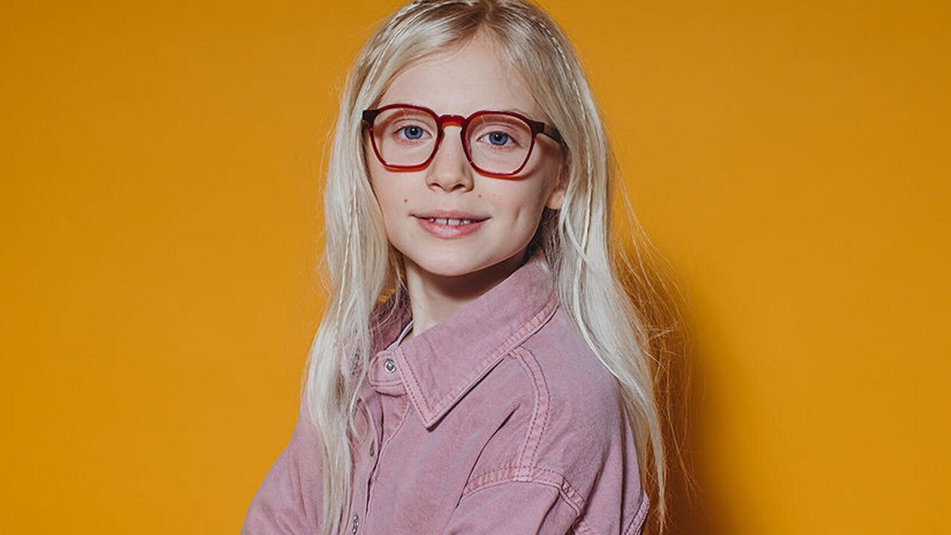 A young child with long blonde hair, wearing red glasses and a light purple shirt, smiles slightly while posing against a solid orange background.