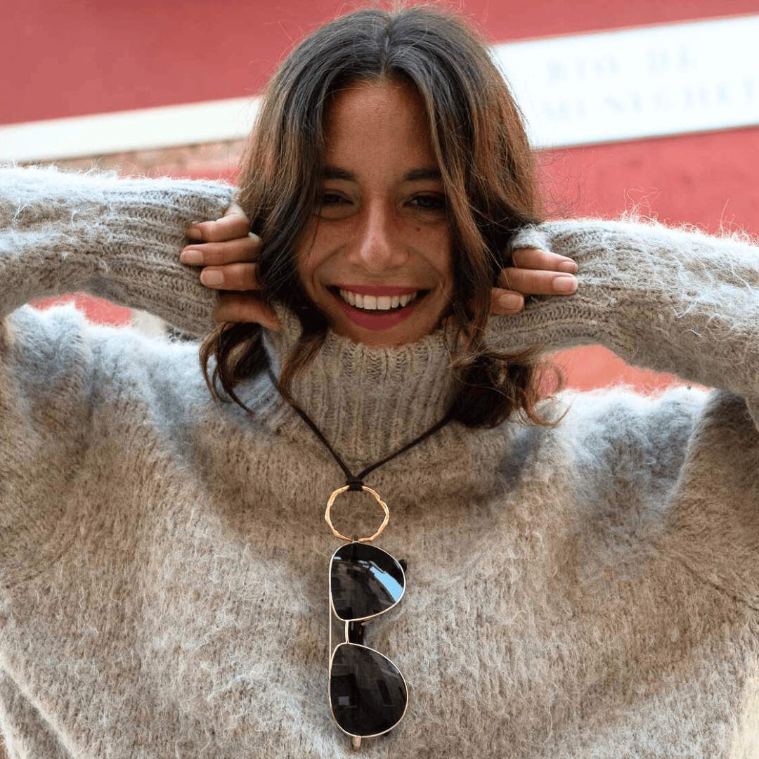 Eine lächelnde Frau in einem flauschigen beigen Pullover zieht den Kragen bis zum Kinn hoch und hält ihn mit beiden Händen fest. Die Sonnenbrille hängt an einem runden Anhänger um ihren Hals. Im Hintergrund ist eine verschwommene rote Wand zu sehen.