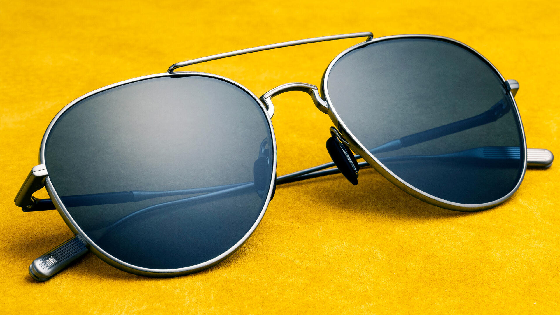 Eine Sonnenbrille im Pilotenstil mit dunkelblauen Gläsern und einem silbernen Metallrahmen liegt auf einer leuchtend gelben Oberfläche.