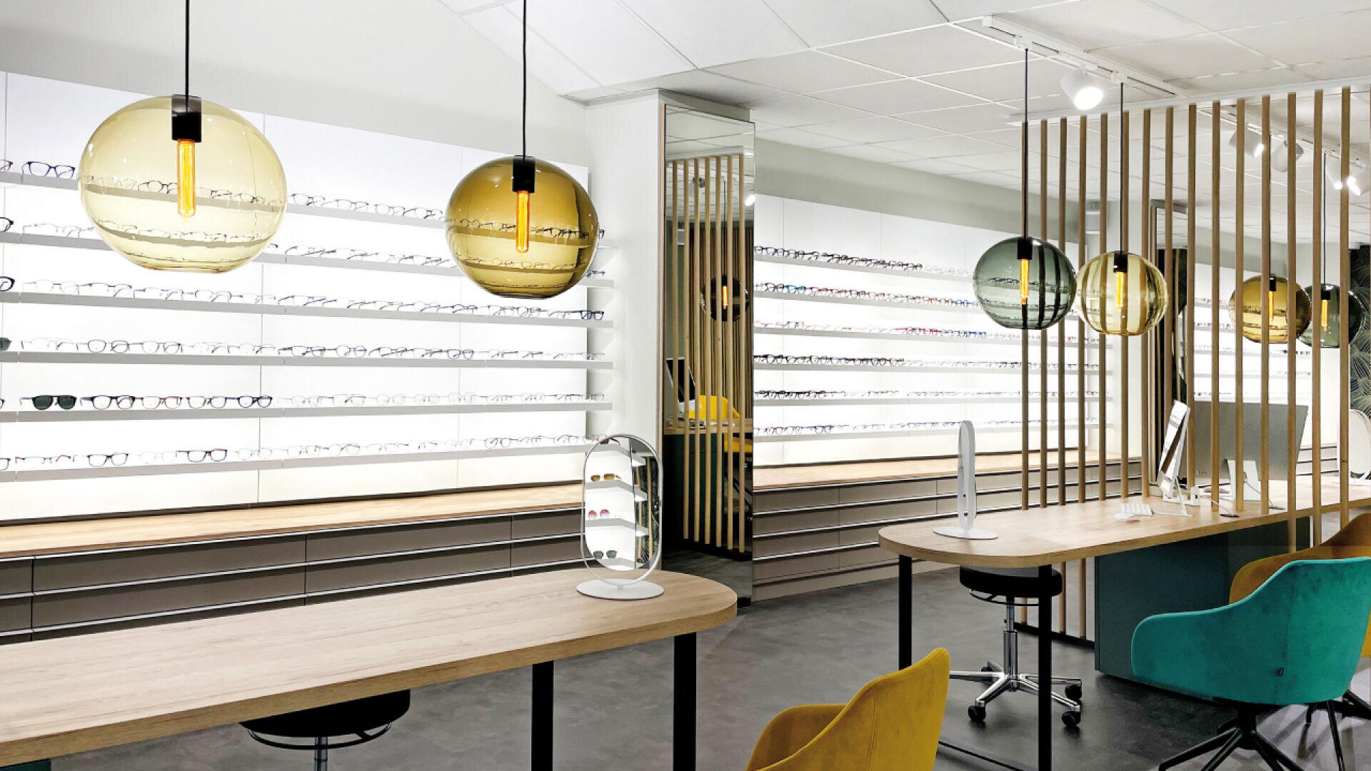 Moderne Optiker-Geschäftseinrichtung mit Brillenreihen auf weißen Regalen, Holztischen mit Stühlen, bunten Glaspendelleuchten und einem Computer auf dem Schreibtisch. Der Raum ist hell und stilvoll.