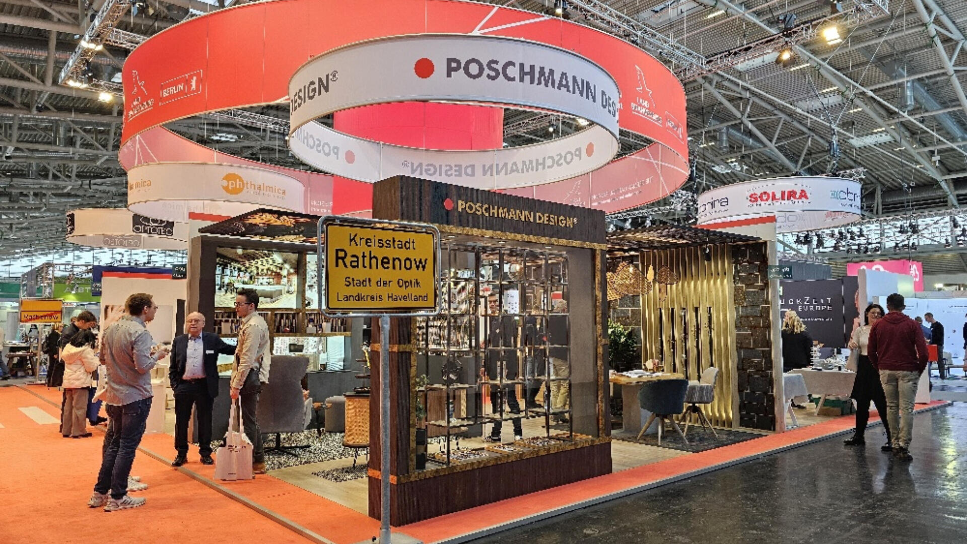 Ein Messestand von POSCHMANN DESIGN ist modern möbliert und dekoriert. In der Nähe unterhalten sich mehrere Personen. Über dem Stand hängen große runde Banner unter einer Decke aus Stahl und Glas.