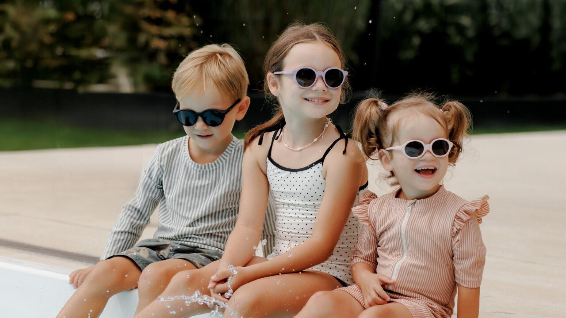 Three young children wearing sunglasses and summer outfits sit on the edge of a pool, smiling and splashing water with their feet on a sunny day.