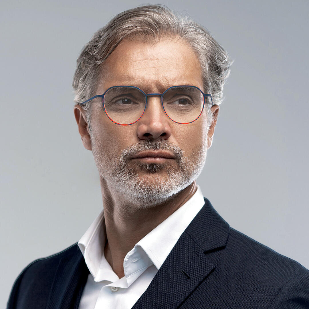 A middle-aged man with gray hair, a short beard, and mustache wears round eyeglasses and a dark suit jacket over a white shirt, looking confidently to the side against a plain background.