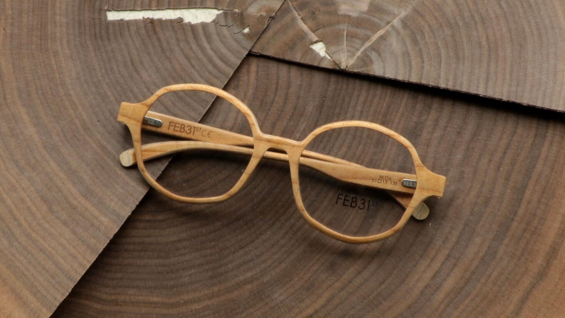 Round, light-colored wooden glasses with engraved FEB31st on the temples, which rest on overlapping pieces of wood with visible annual rings and natural structures.