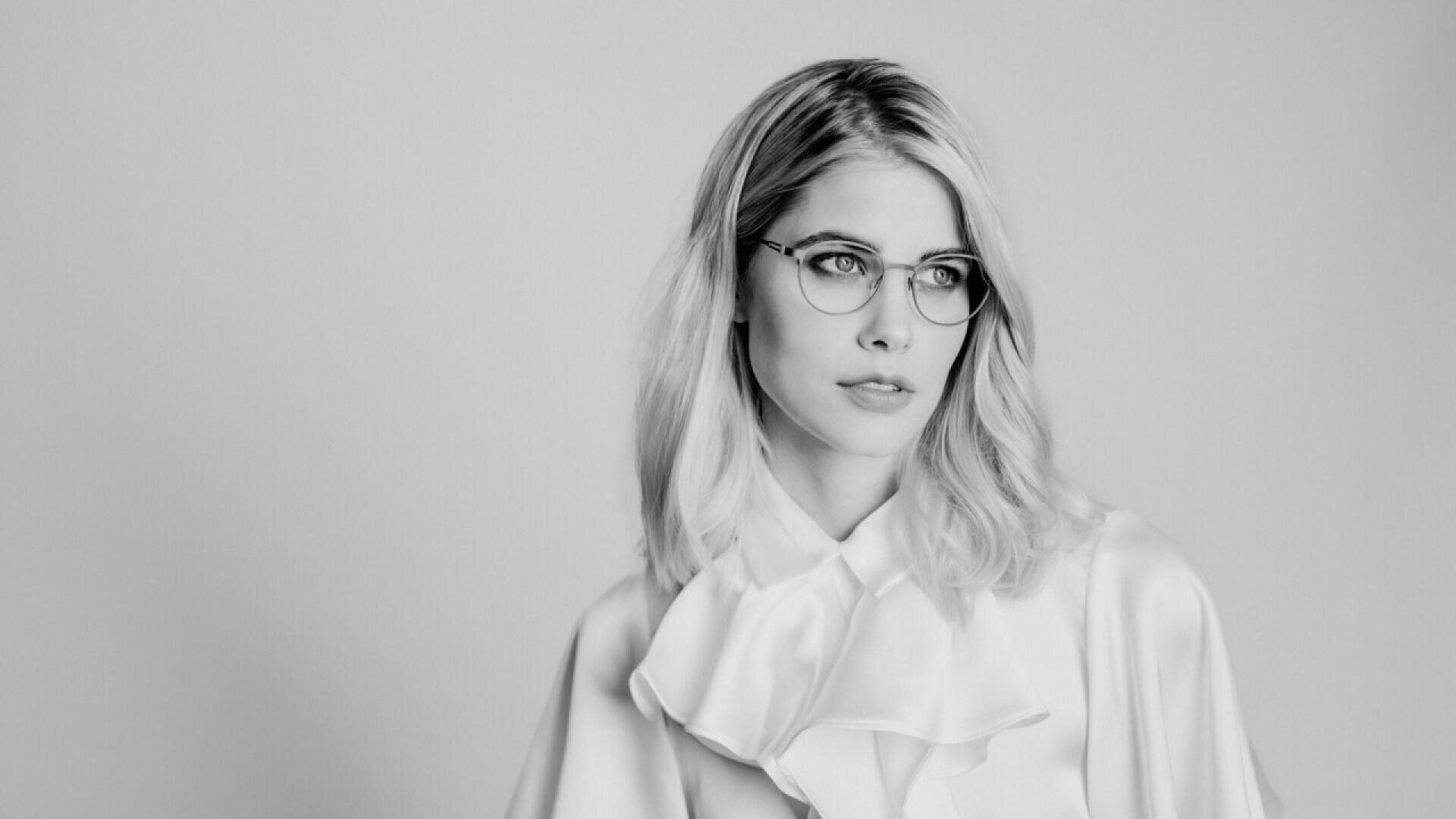 A black and white photo of a woman with shoulder-length light hair wearing glasses and a blouse with ruffles, looking slightly to the side against a plain background.