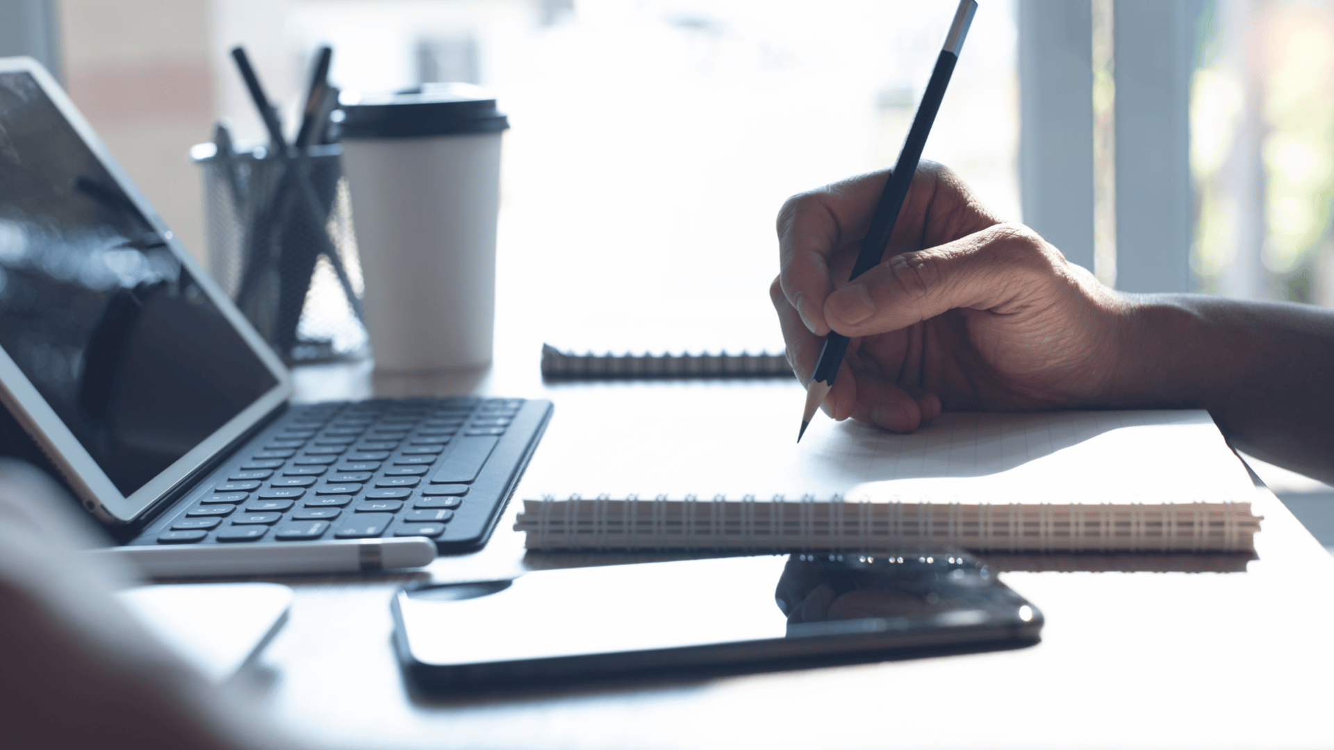 Eine Person schreibt mit einem Bleistift in ein Notizbuch an einem Schreibtisch, daneben ein Tablet mit Tastatur, ein Smartphone, eine Kaffeetasse und ein Stifthalter in einem hellen, modernen Arbeitsbereich.