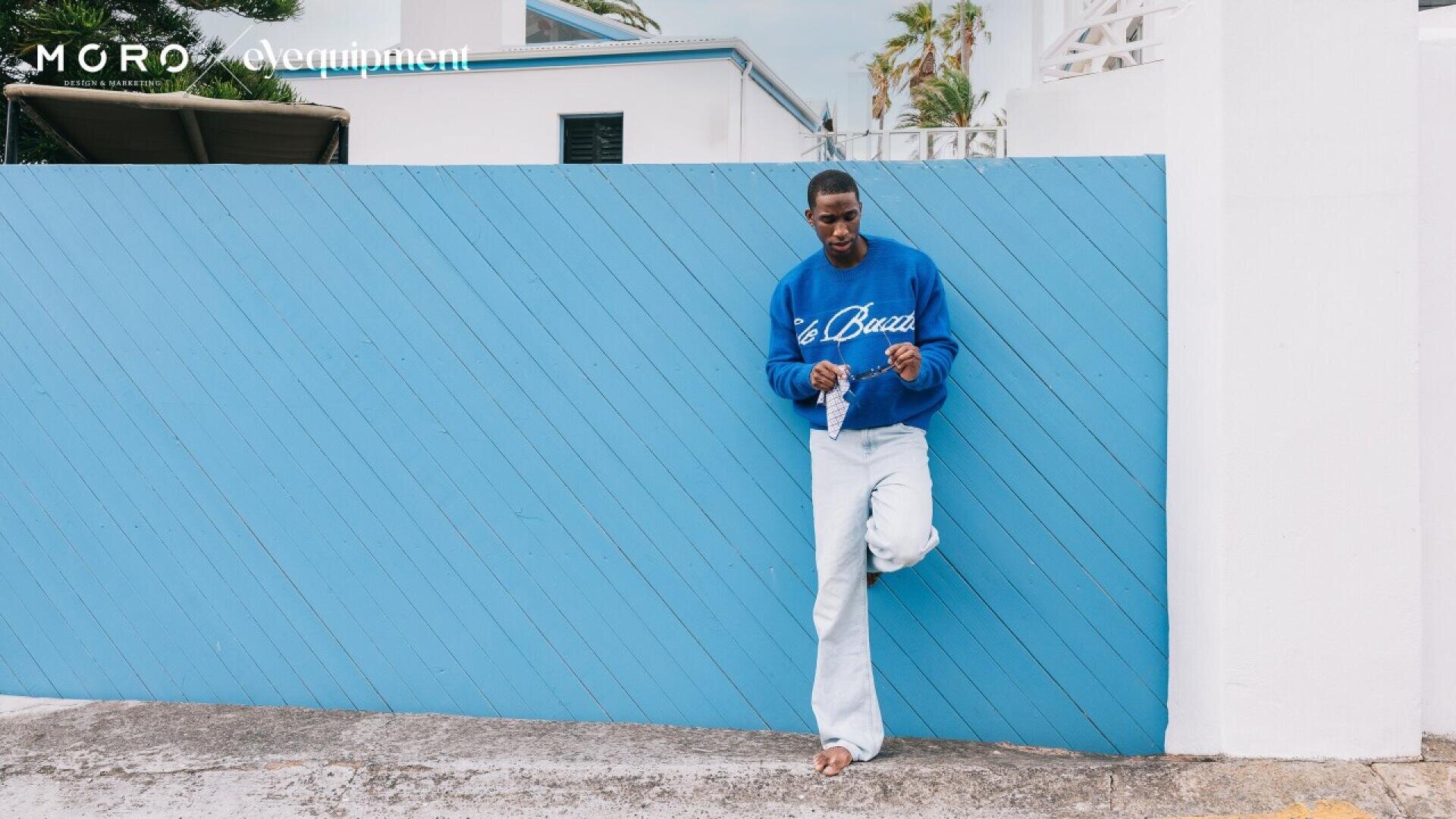 A man wearing a blue sweatshirt and light jeans leans against a light blue wooden fence outdoors, looking down and holding something in his hands.