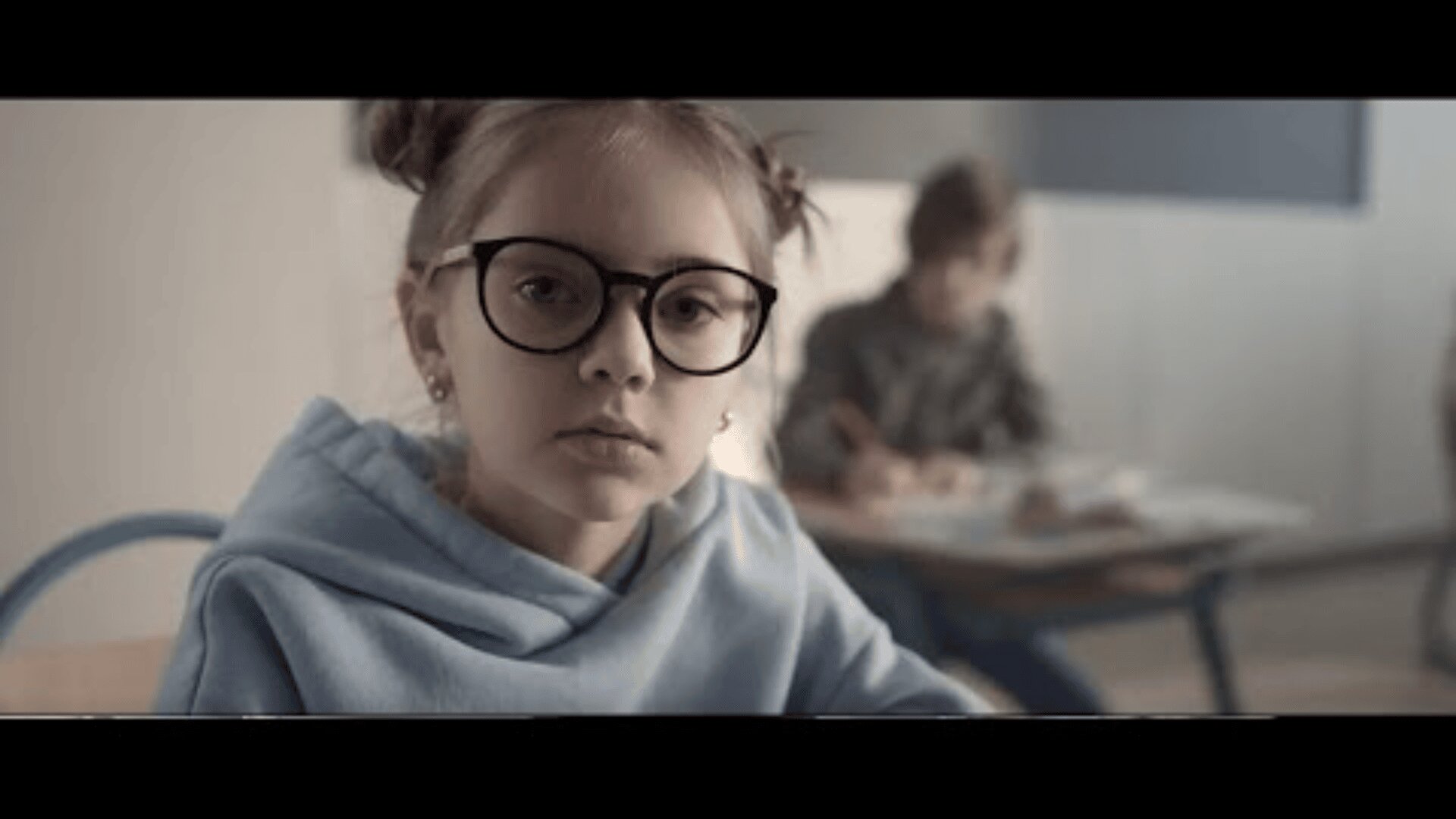 Ein junges Mädchen mit Brille und hellblauem Kapuzenpullover sitzt in einem Klassenzimmer an einem Schreibtisch und blickt in die Kamera. Im Hintergrund schreibt ein weiteres Kind an einem Schreibtisch. Die Szene ist in sanften, neutralen Farbtönen gehalten.