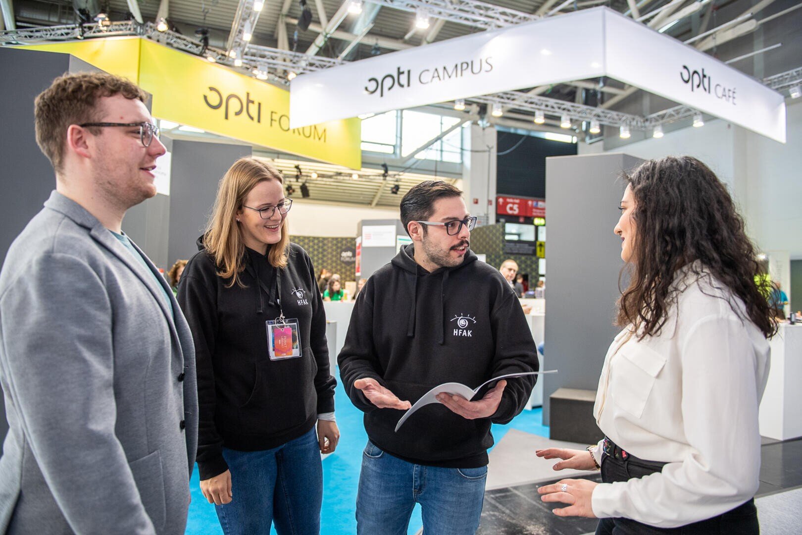 Four people are in conversation at an indoor trade fair or conference. Two of them are wearing black hoodies with a logo, one is holding an open magazine, another is wearing a badge. Trade fair stands and signs with the word "opti" can be seen in the background.