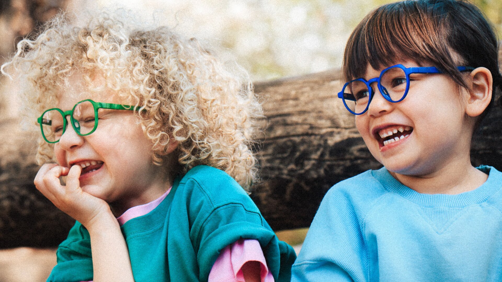 Zwei Kinder mit bunten Brillen und blauen Hemden sitzen im Freien und lächeln und lachen. Das eine hat lockiges blondes Haar und eine grüne Brille, das andere hat glattes braunes Haar und eine blaue Brille. Im Hintergrund ist ein Baumstamm zu sehen.