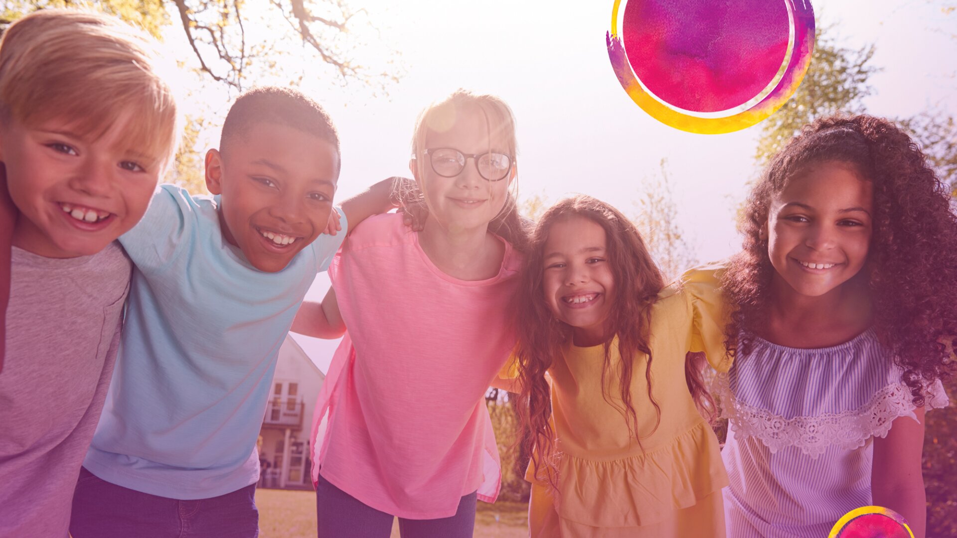 Fünf lächelnde Kinder stehen im Freien unter hellem Sonnenlicht dicht beieinander, die Arme umeinander gelegt, mit Bäumen im Hintergrund. Das Bild hat bunte Kreise in der oberen rechten Ecke zur Dekoration.