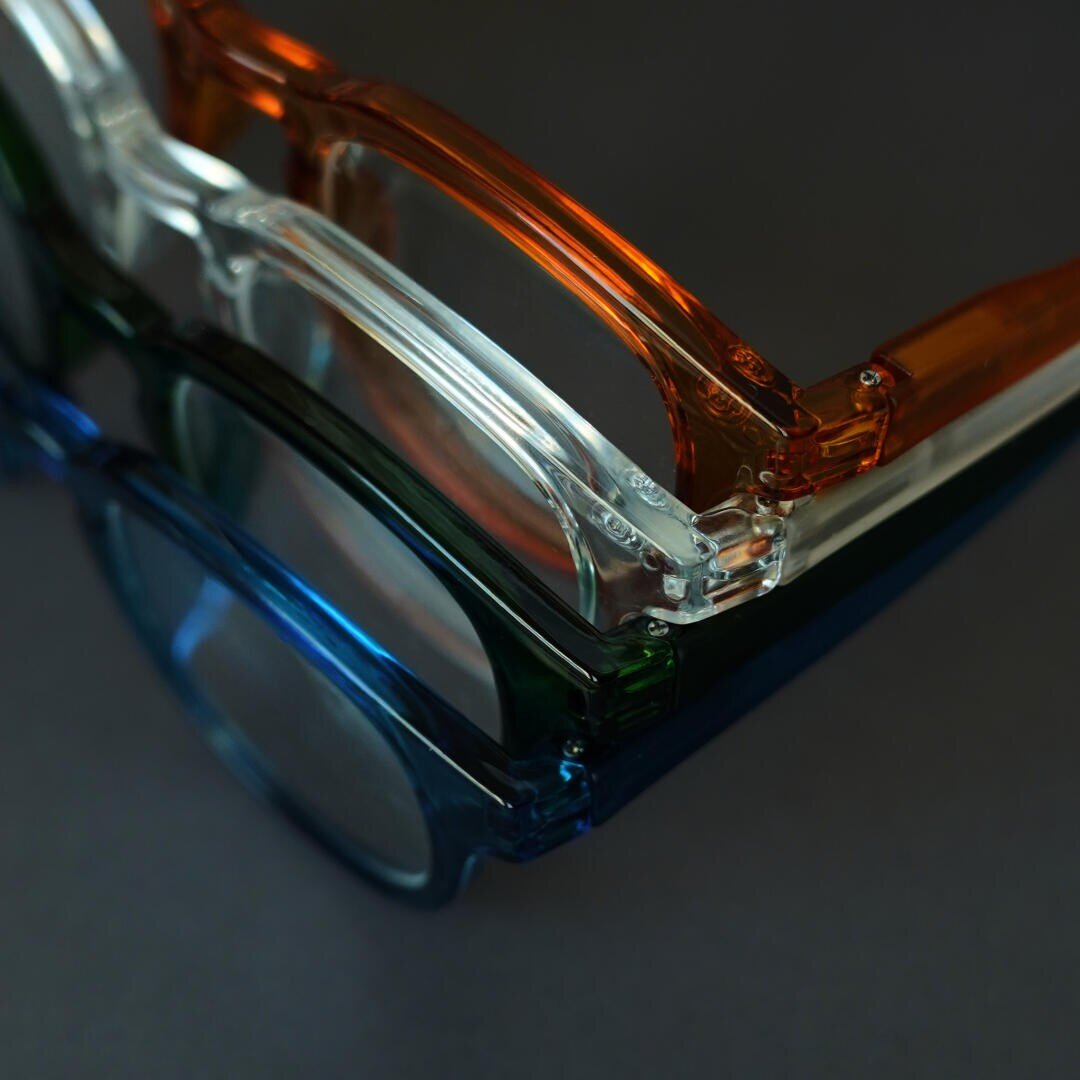Three pairs of glasses with transparent frames - orange, clear and blue - are stacked vertically on a dark surface and viewed up close.