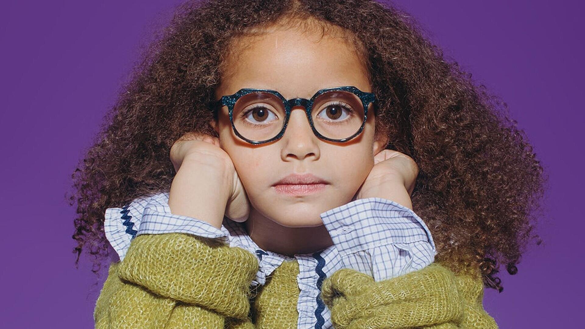 A young child with curly hair, wearing round glasses, a green sweater, and a blue checkered shirt, rests their chin on their hands against a solid purple background.