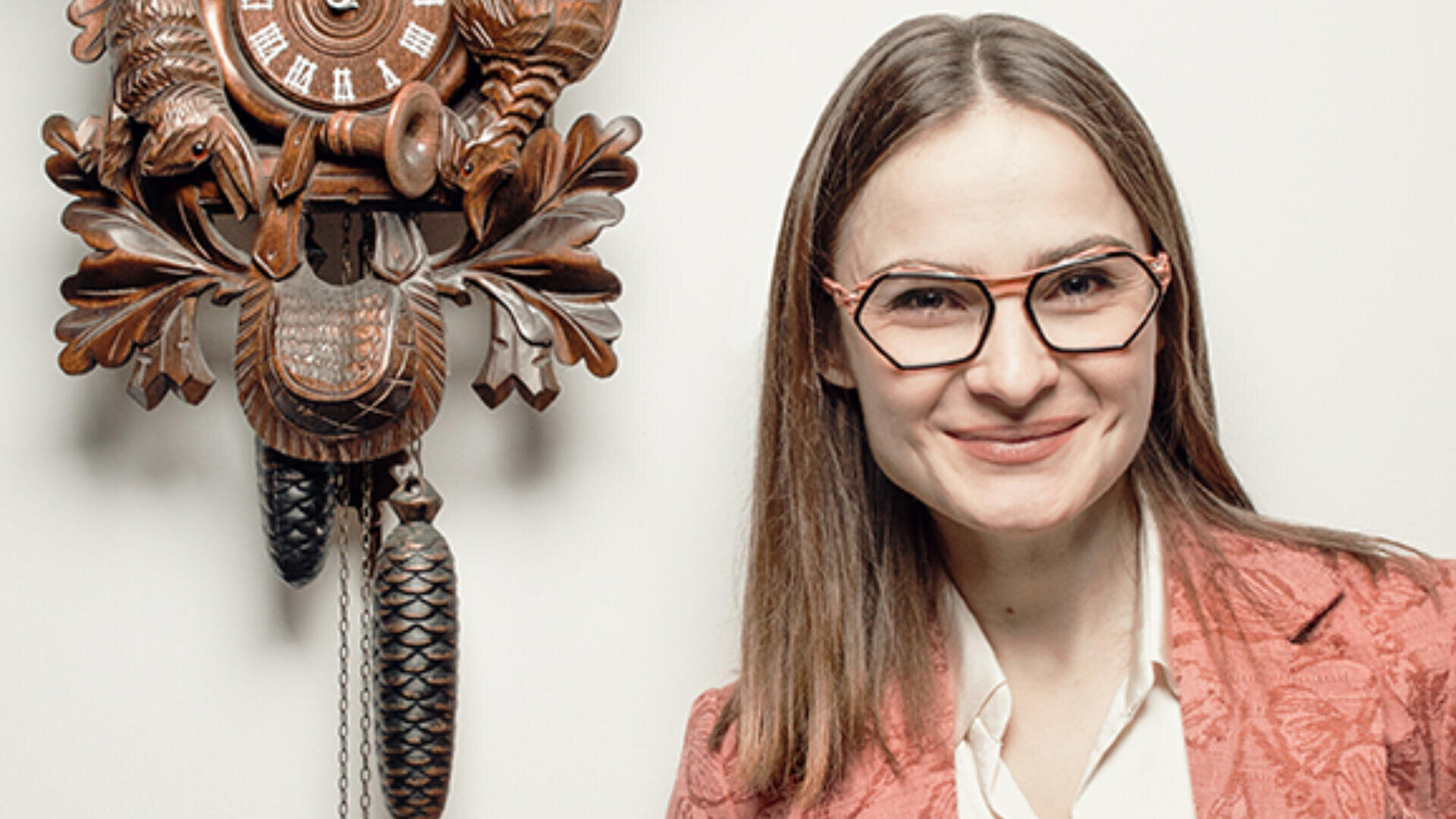 Eine Frau mit glattem braunem Haar und geometrischer Brille steht lächelnd neben einer dekorativen hölzernen Kuckucksuhr an einer weißen Wand. Sie trägt einen rosa gemusterten Blazer über einem weißen Hemd.