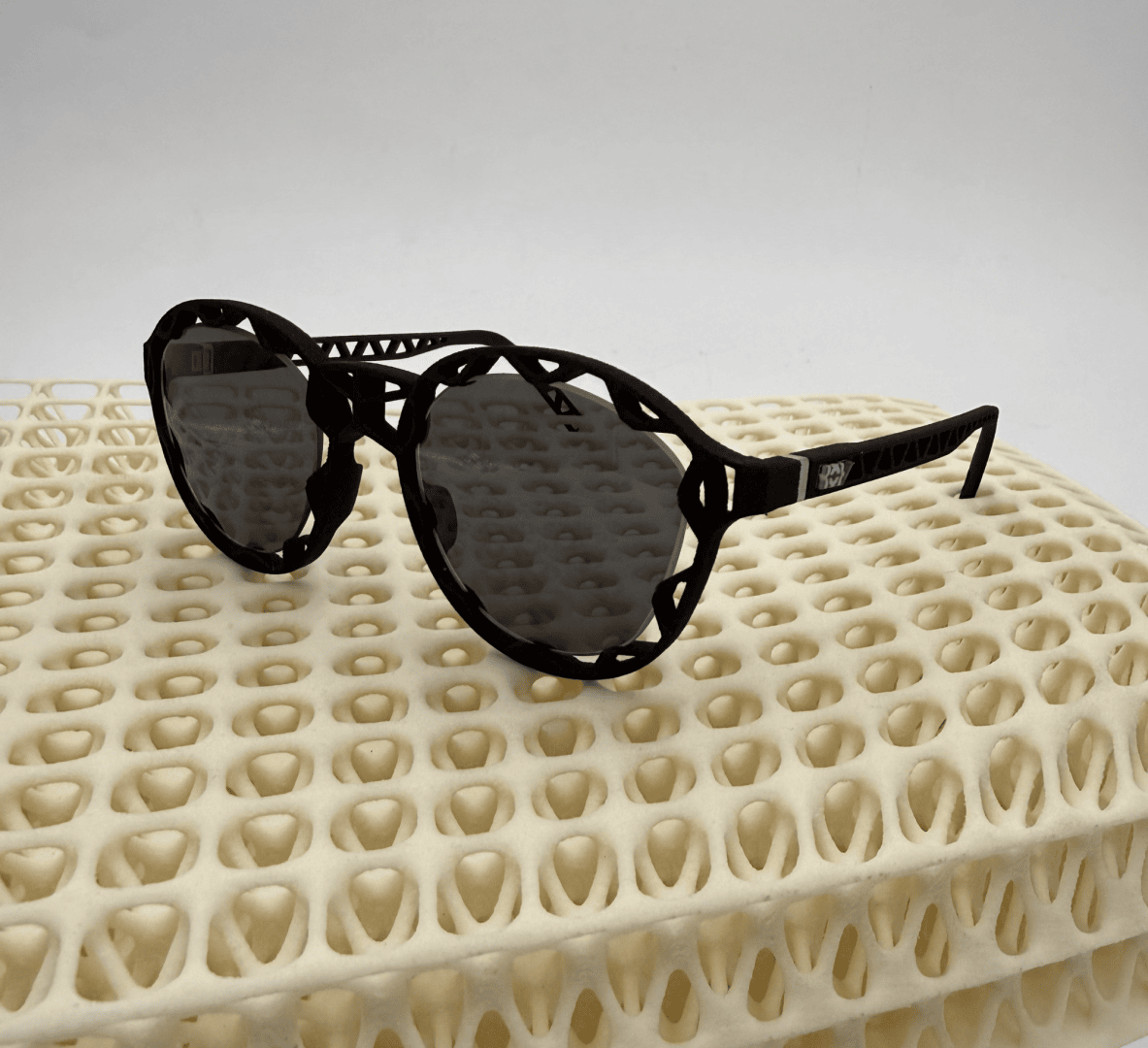 Round black sunglasses with a decorative, wavy frame rest on a cream-colored, grid-like patterned surface against a plain white background.
