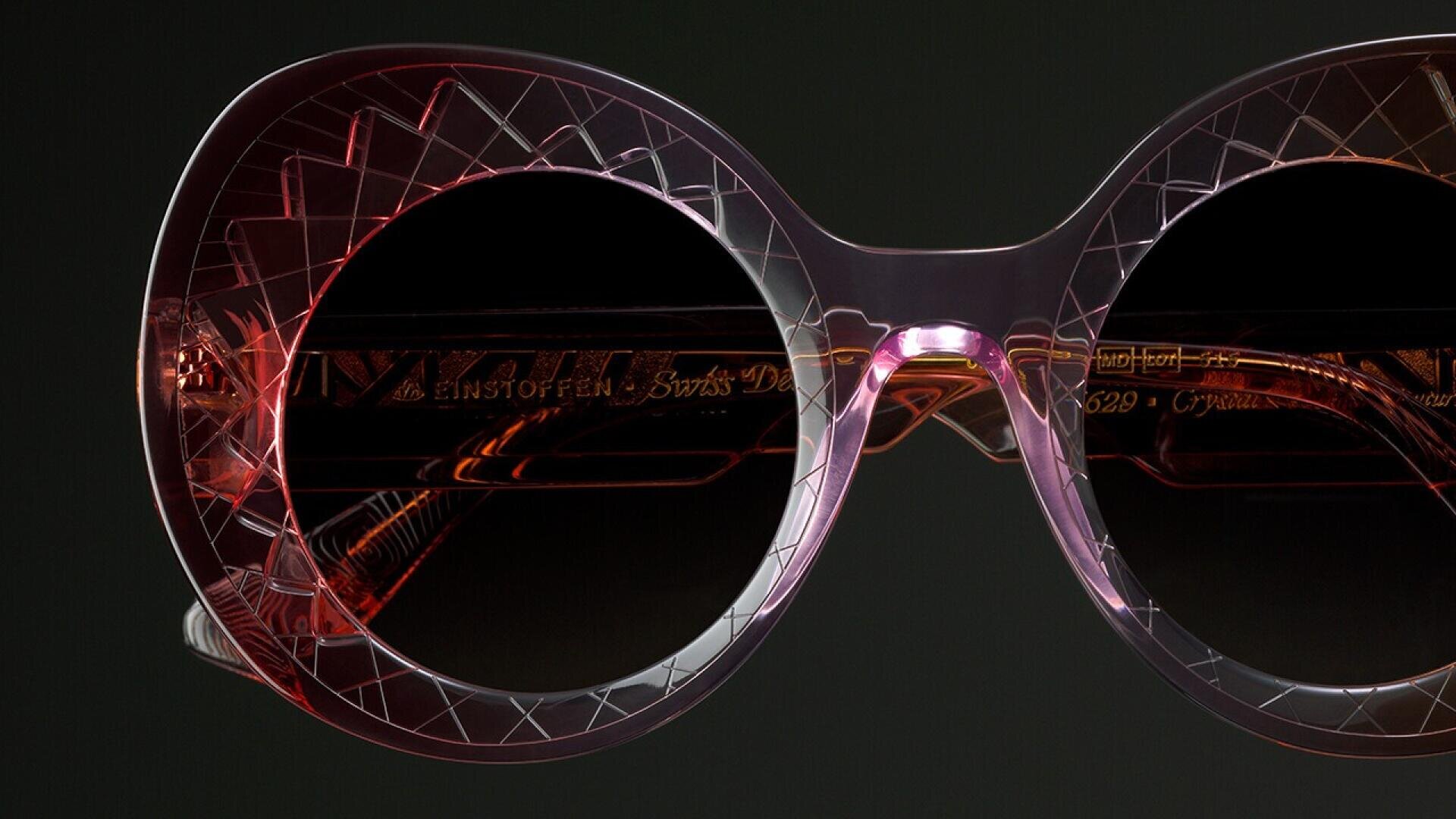Close-up of stylish, round sunglasses with translucent pink frames featuring a geometric etched pattern and dark lenses against a black background.