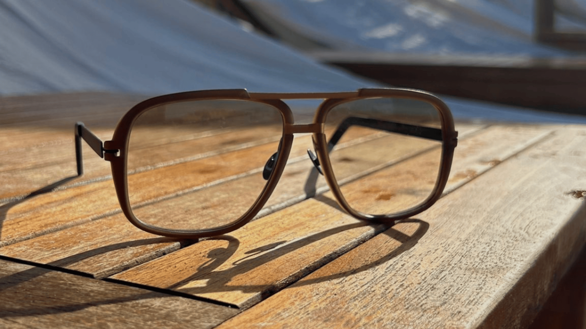 Eine braune Pilotenbrille liegt im Sonnenlicht auf einer Holzoberfläche und wirft einen deutlichen Schatten. Im unscharfen Hintergrund ist weiße Leinwand oder Stoff zu sehen.