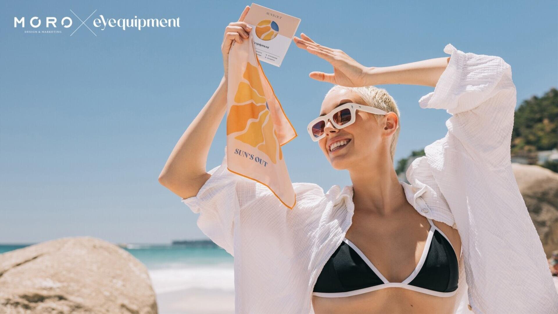 A smiling person in sunglasses and a white shirt over a black bikini holds up a colorful cloth and card on a sunny beach, with rocks and ocean in the background. Text reads MGRO x eyequipment in the top left corner.