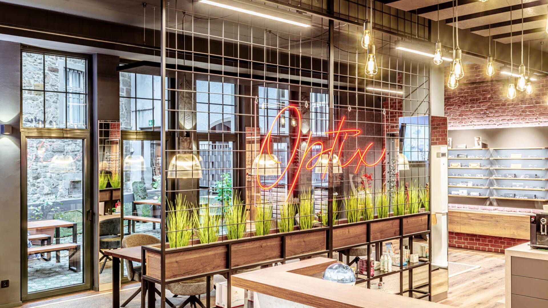 Modernes Interieur eines Cafés oder Büroraums mit großen Fenstern, unverputzten Ziegelwänden, Hängelampen und einem Optix-Neonschild. Grüne Pflanzen sind auf einer hölzernen Trennwand mit Metallgitter angeordnet.