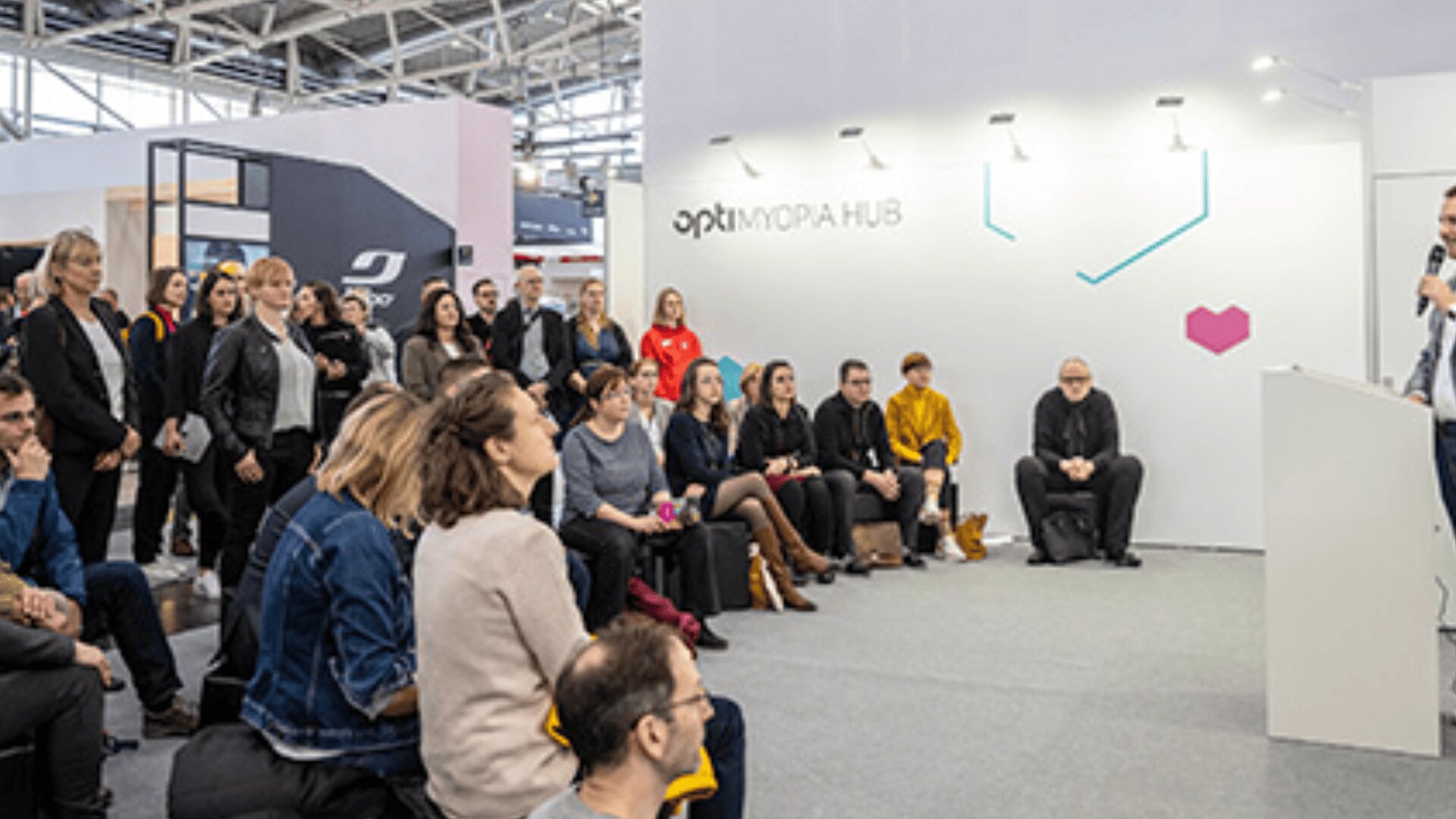 Eine Gruppe von Menschen sitzt und steht aufmerksam um einen Redner bei einer modernen Indoor-Veranstaltung oder Konferenz, mit weißen Wänden und der Aufschrift optimum FORA HUB im Hintergrund.