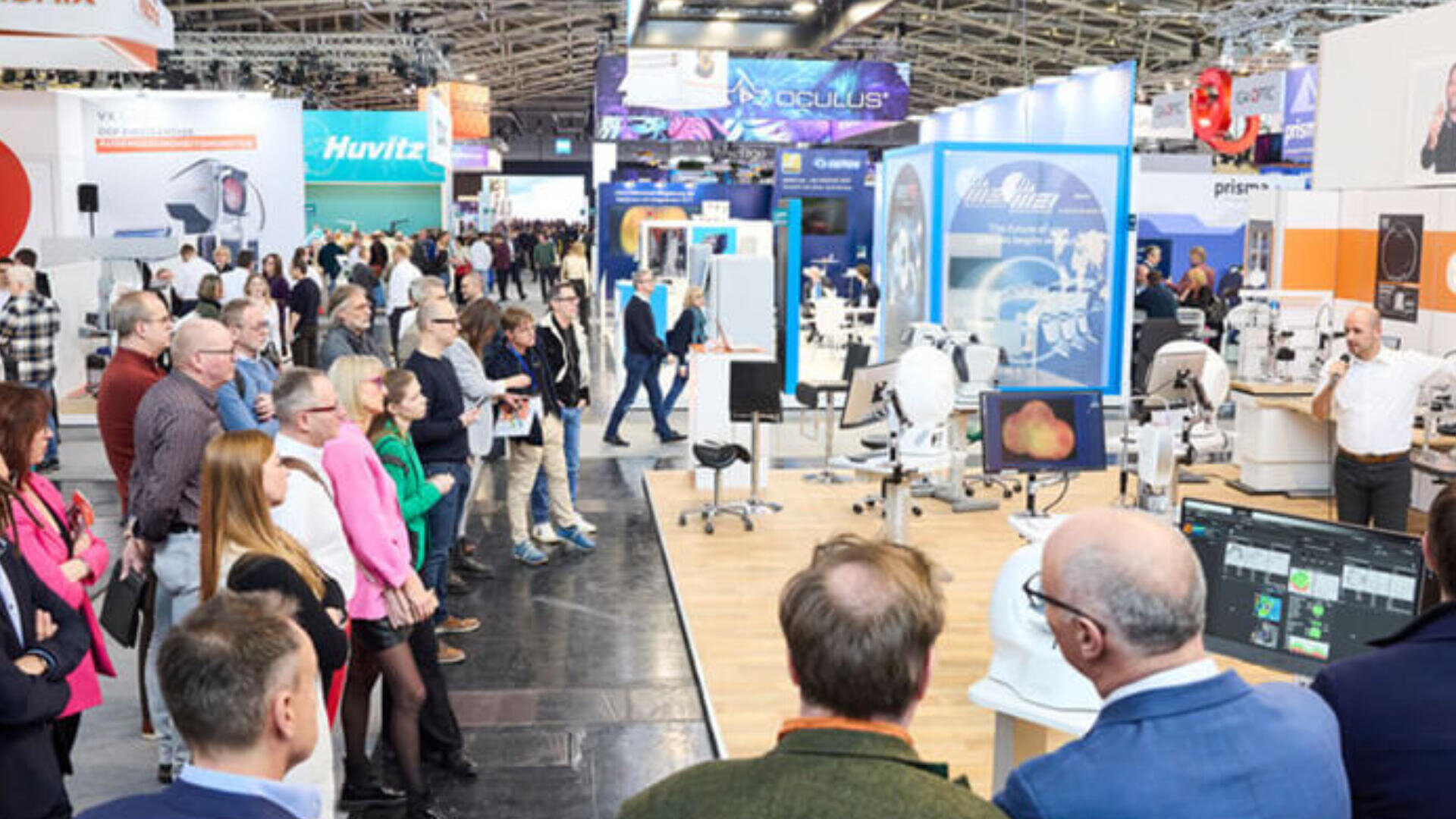 Eine Gruppe von Menschen steht auf einer geschäftigen Handelsmesse oder Ausstellung und hört einem Vortragenden zu. Sie ist umgeben von Ständen mit medizinischen oder technischen Geräten und Informationsbildschirmen.