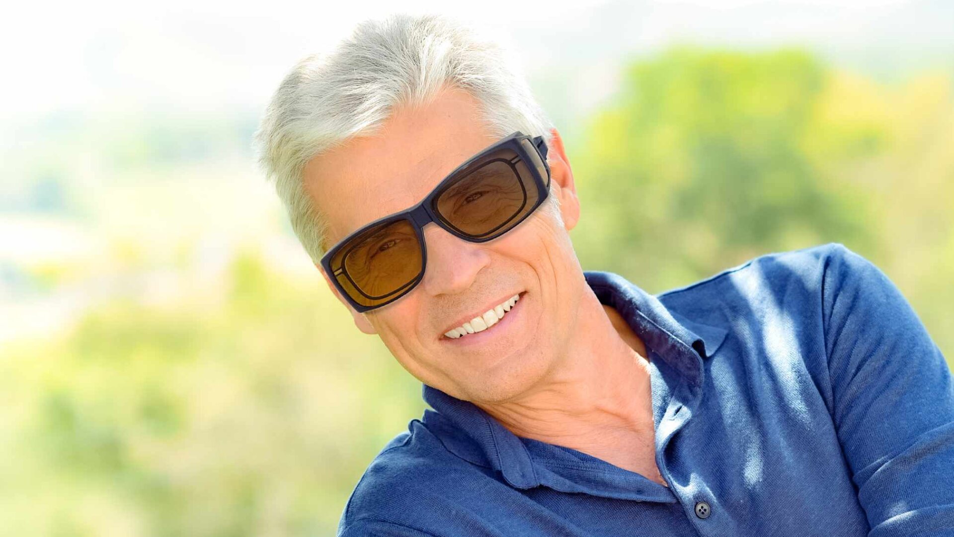 Lächelnder älterer Mann mit kurzen grauen Haaren, der eine große dunkle Sonnenbrille und ein blaues Hemd mit Kragen trägt, steht im Freien vor einem verschwommenen grünen und gelben Hintergrund.