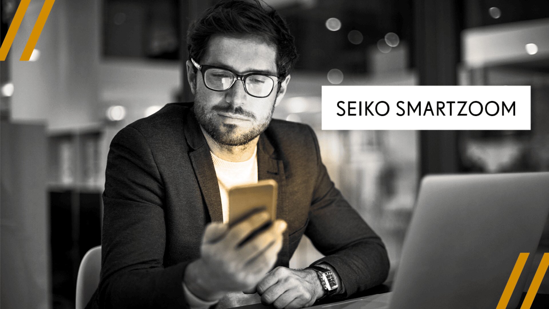 Ein Mann mit Brille und Anzug sitzt mit einem Laptop an einem Schreibtisch und schaut auf sein Smartphone. Auf der rechten Bildseite erscheint der Schriftzug SEIKO SMARTZOOM.