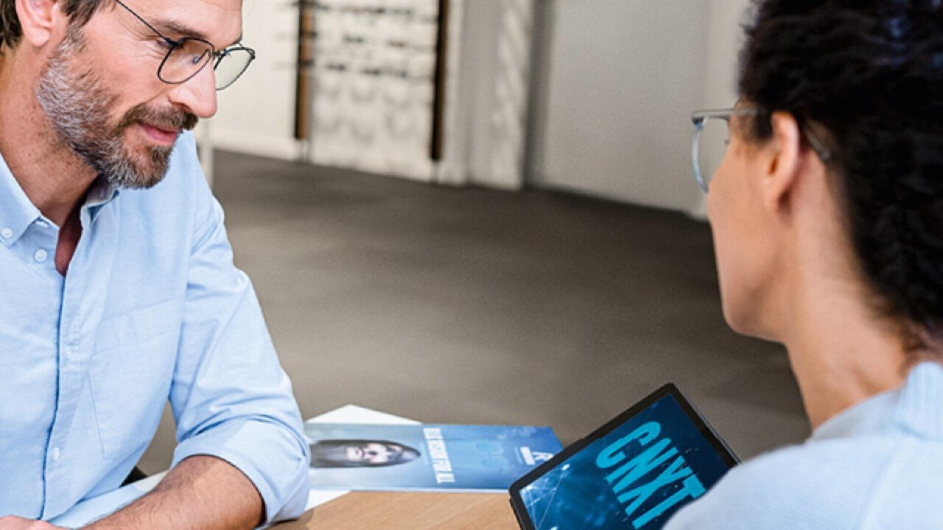 A man and a woman, both wearing glasses, sit at a table having a discussion. The woman holds a tablet displaying the word CNXT, and a brochure with a womans face is visible on the table.
