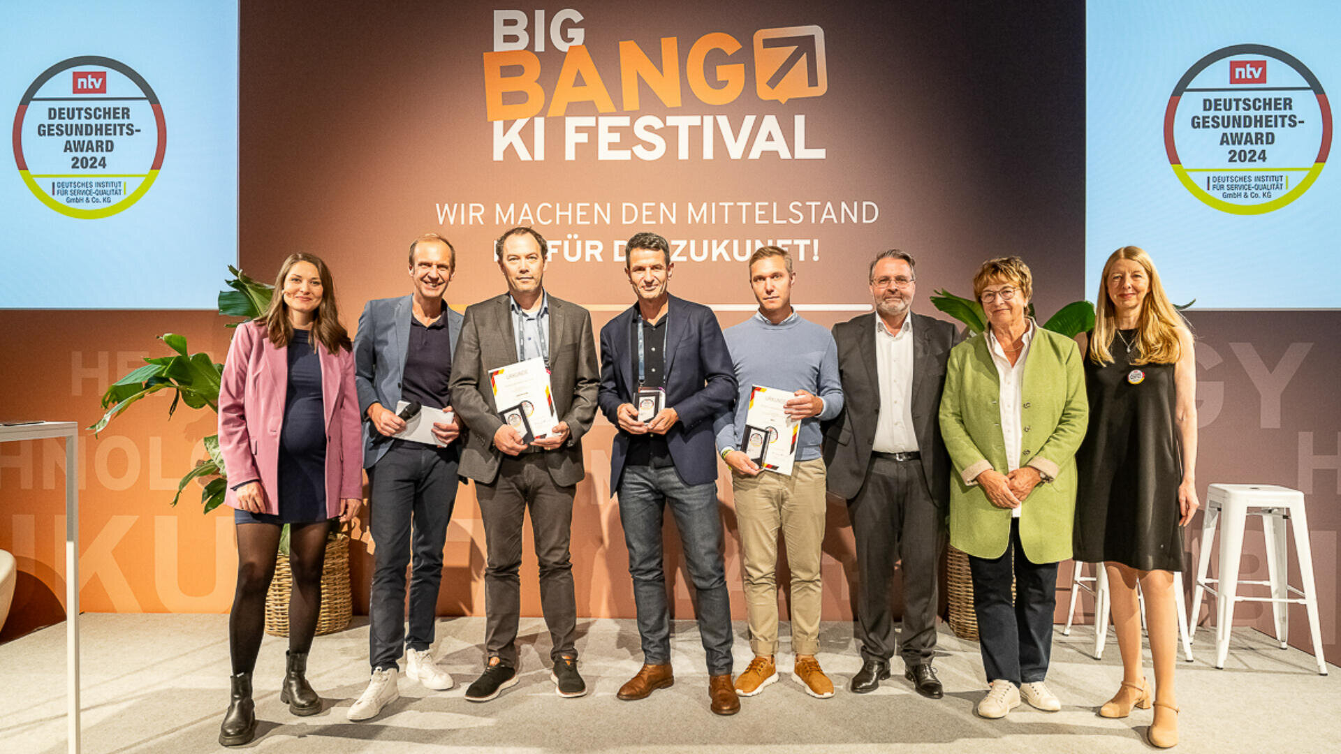 Acht Personen stehen beim Big Bang KI Festival auf der Bühne, einige halten Auszeichnungen und Urkunden in den Händen. Dahinter hängt ein Banner mit dem Veranstaltungsnamen und den Logos des Deutschen Gesundheits-Award 2024.