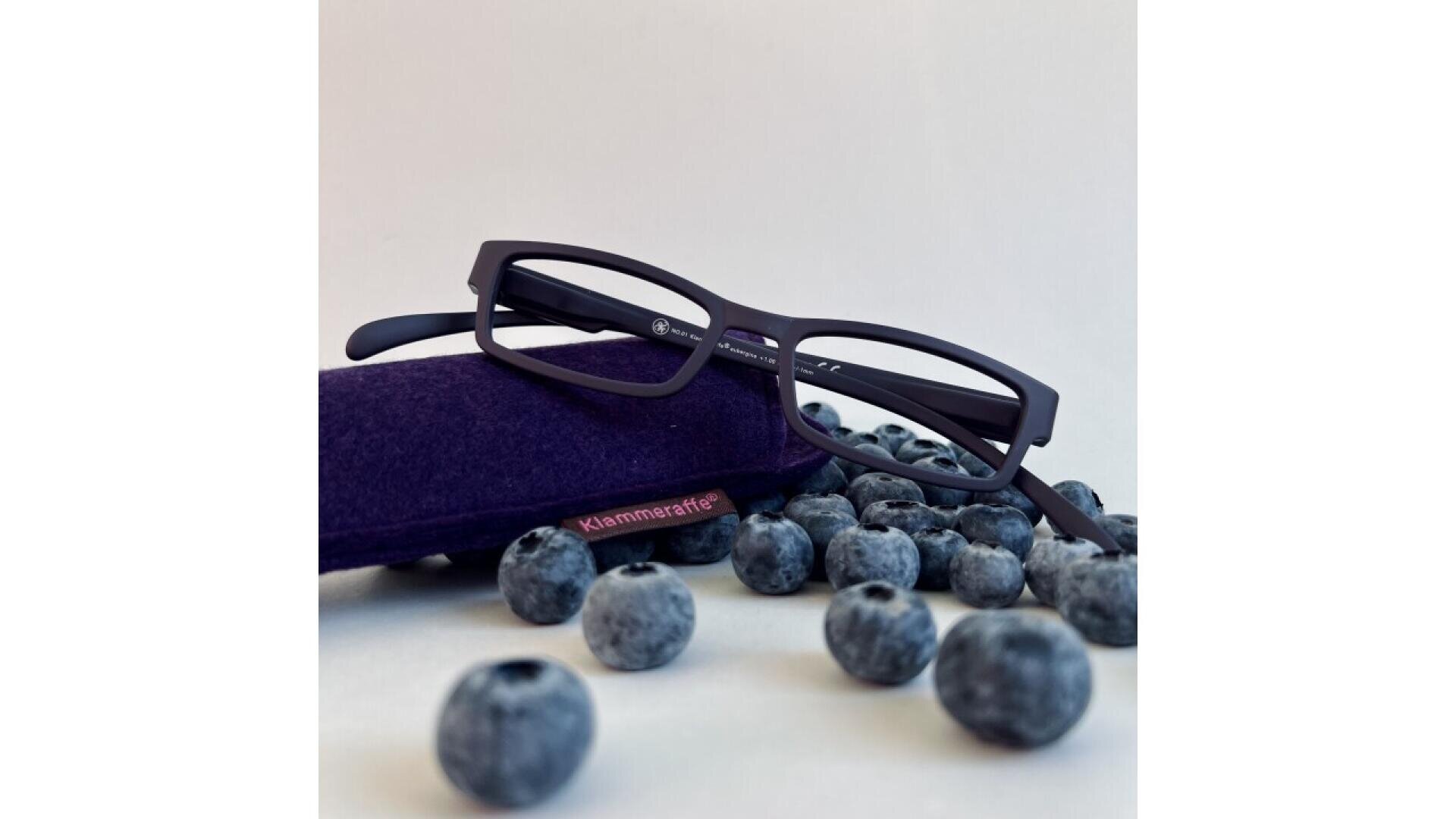 A pair of black eyeglasses rests on a dark purple case surrounded by scattered blueberries against a plain white background.
