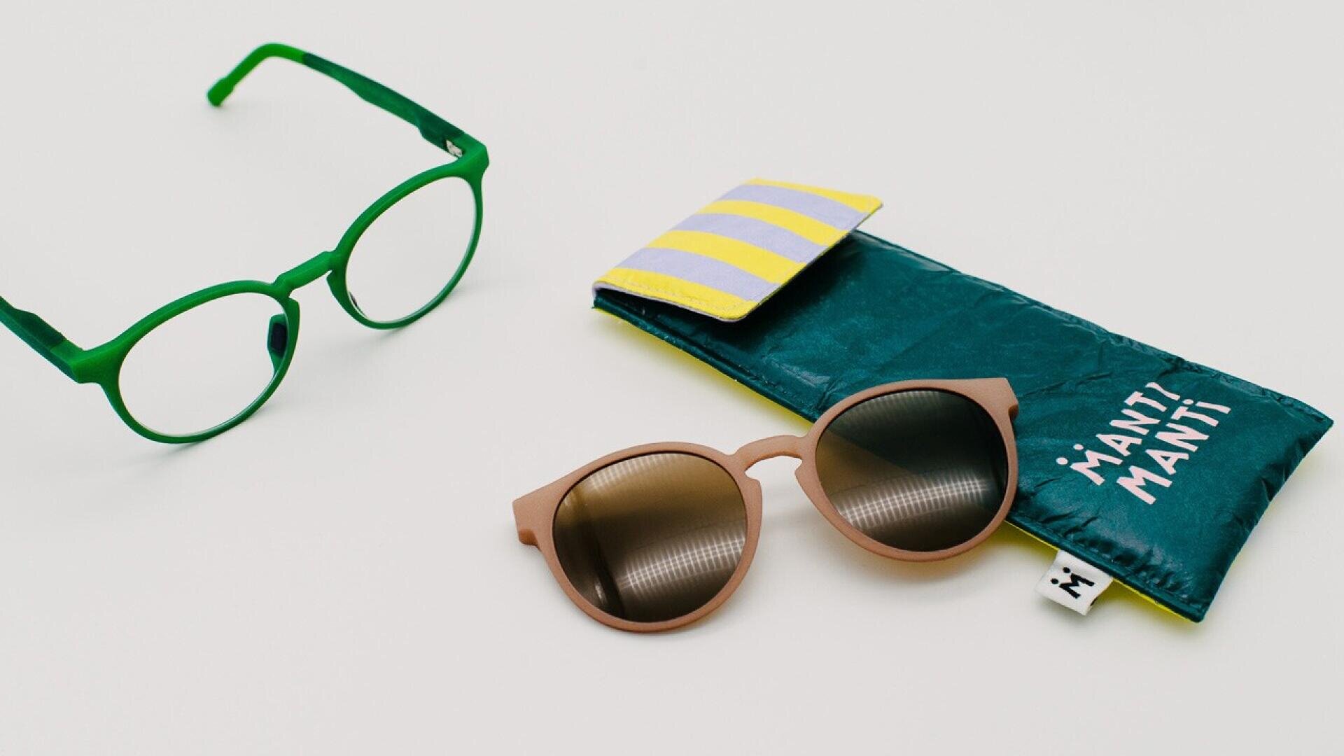 A pair of green round eyeglasses, brown round sunglasses with dark lenses, and a turquoise glasses case with a yellow-striped flap are displayed on a white surface.