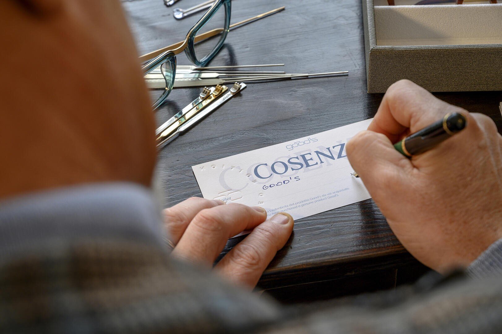 A person in a checked jacket is writing on a card with the inscription COSENZA at a desk. Glasses, tools and an open box can be seen nearby.