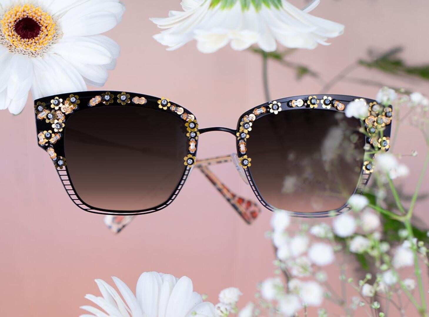 Eine Cat-Eye-Sonnenbrille mit schwarzem Rahmen, der mit kleinen Blumendekorationen verziert ist, umgeben von weißen Blumen und vor einem zartrosa Hintergrund.