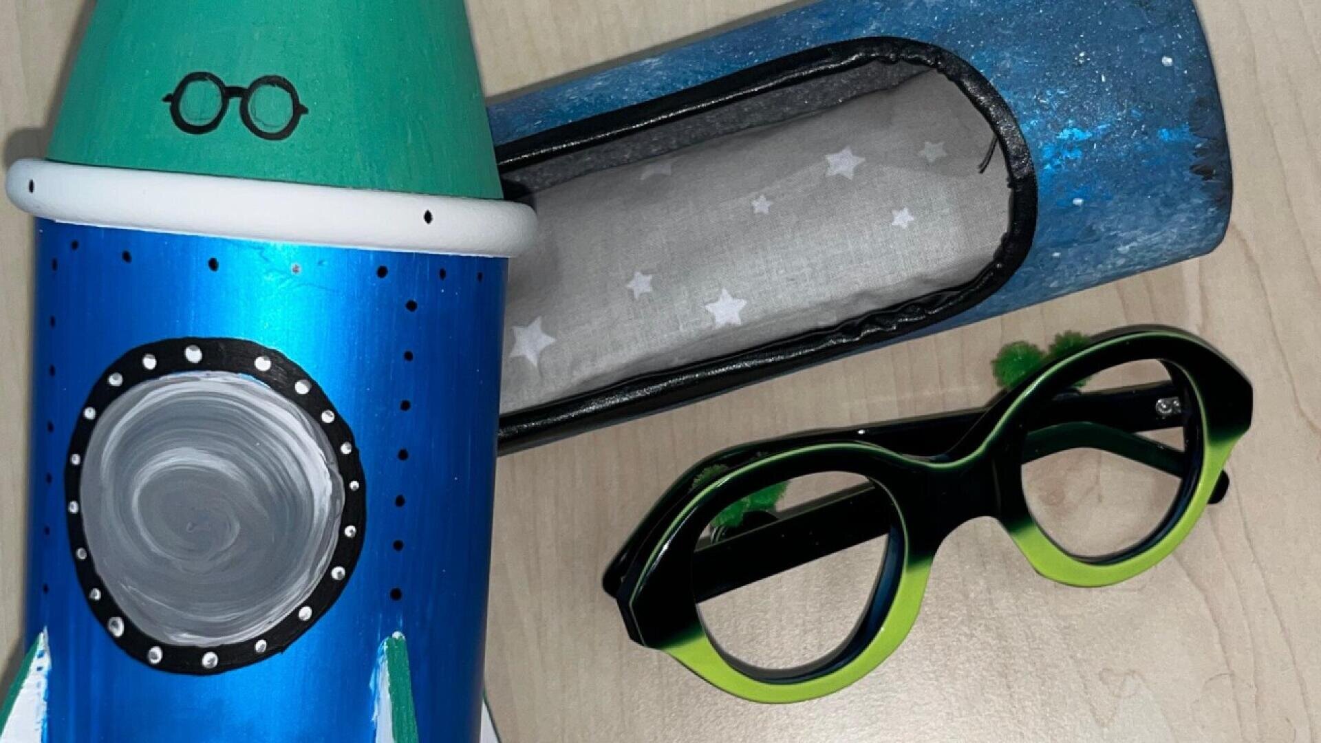 A painted blue and green rocket ship with a porthole and cartoon glasses is next to a black and green pair of glasses and a blue rectangular case with a star-patterned lining, all on a light wooden surface.
