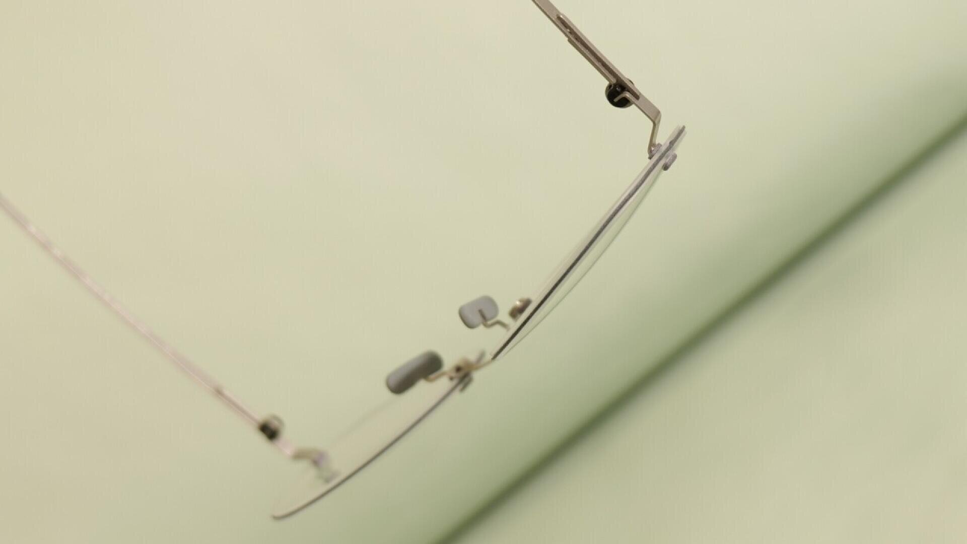 A close-up of a pair of eyeglasses with thin metal frames, focusing on the nose pads and rim against a blurred light green background.