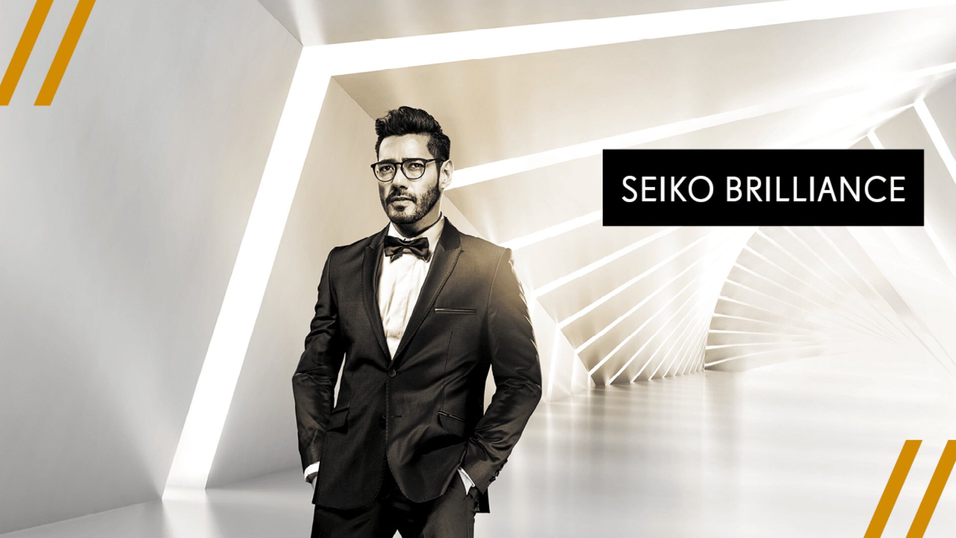 Ein Mann in einem schwarzen Smoking steht selbstbewusst in einem modernen, weißen, geometrischen Flur. Neben ihm erscheint der Text SEIKO BRILLIANCE. Drei goldene diagonale Linien zieren die linke obere und die linke untere Ecke.