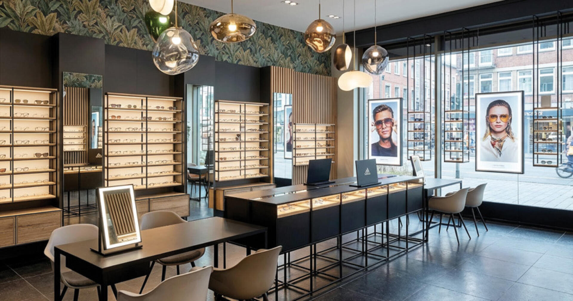 A modern eyewear store with illuminated eyewear shelves, stylish pendant lights, tables with chairs and large advertising posters of spectacle wearers in the shop window.
