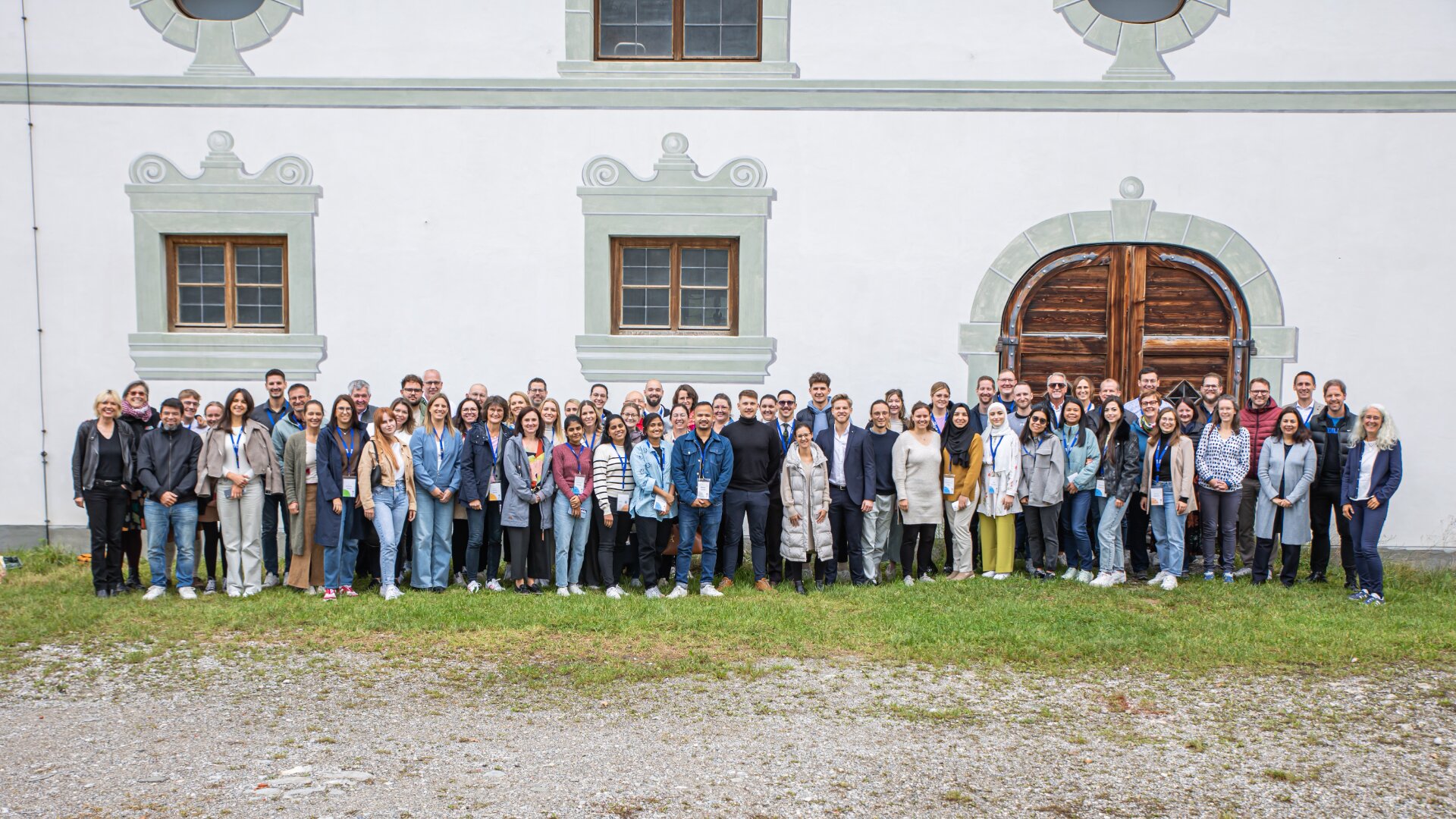 Eine große Gruppe von Menschen steht im Freien vor einem historischen weißen Gebäude mit gewölbten Holztüren und dekorativen Fenstern und posiert für ein Gruppenfoto. Das Wetter ist klar und die Gruppe wirkt fröhlich.