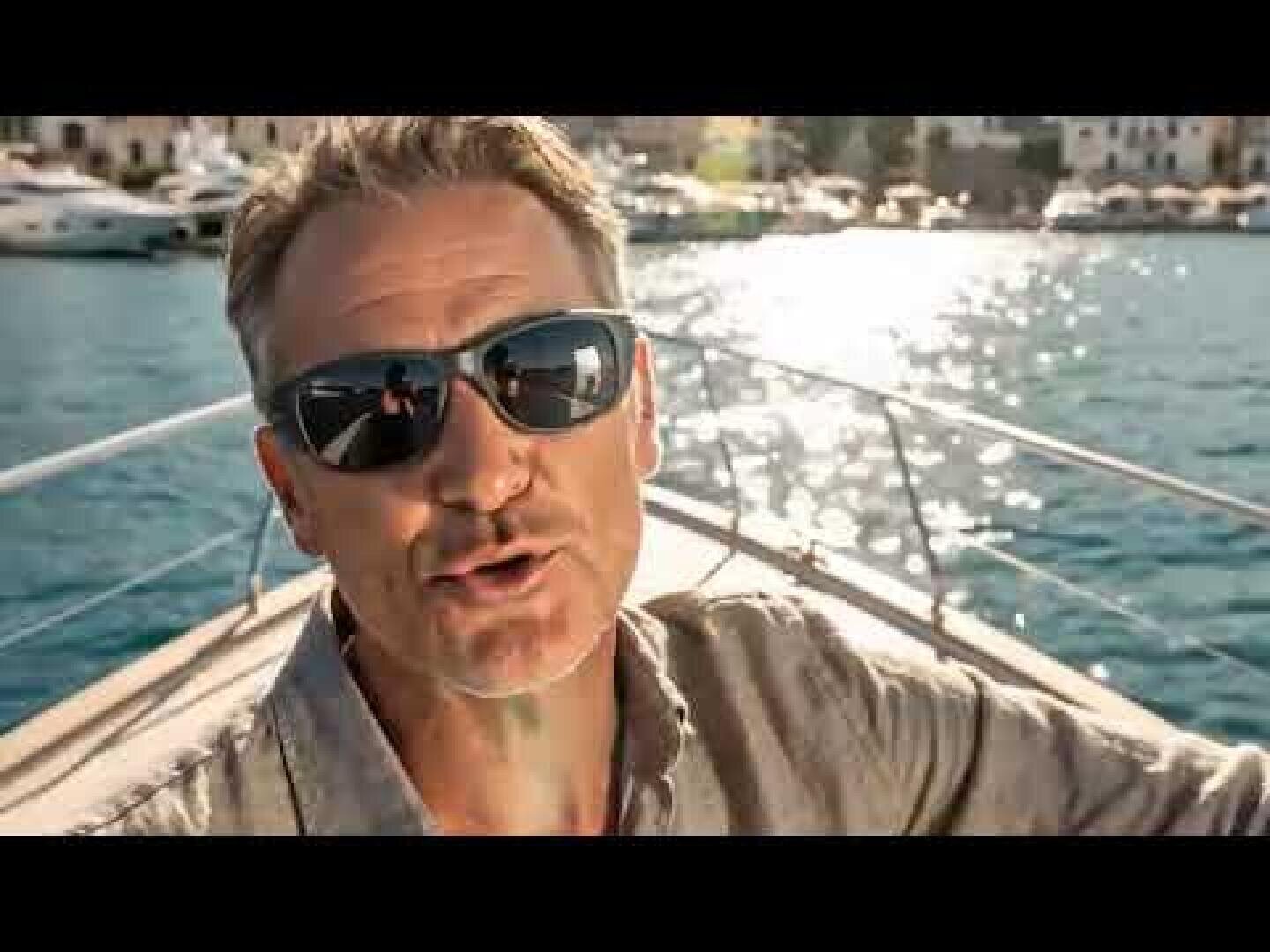 A man with side protection sunglasses and a light-colored shirt sits on a boat and smiles at the camera. The sun is reflected on the water and boats and buildings can be seen in the background.