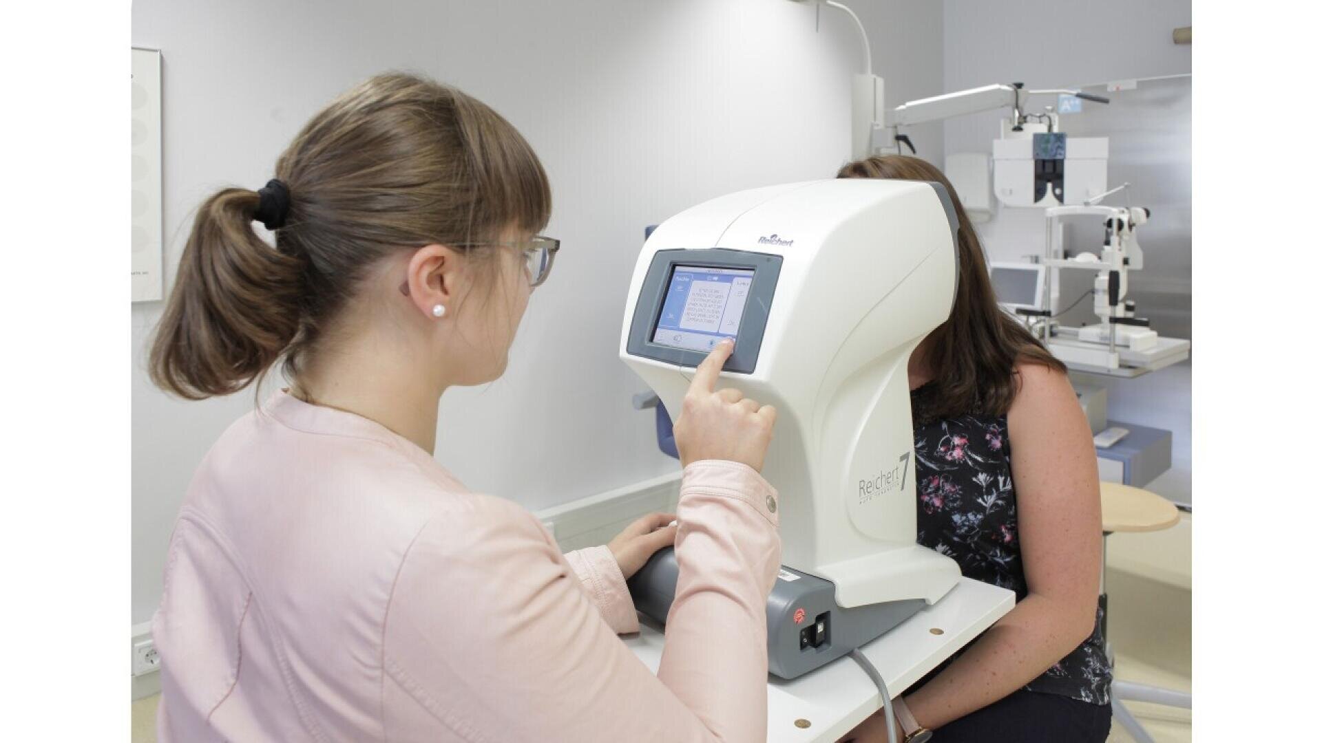 A woman operates an eye examination machine while another woman sits in front of it, partially obscured by the device, in a clinical setting.