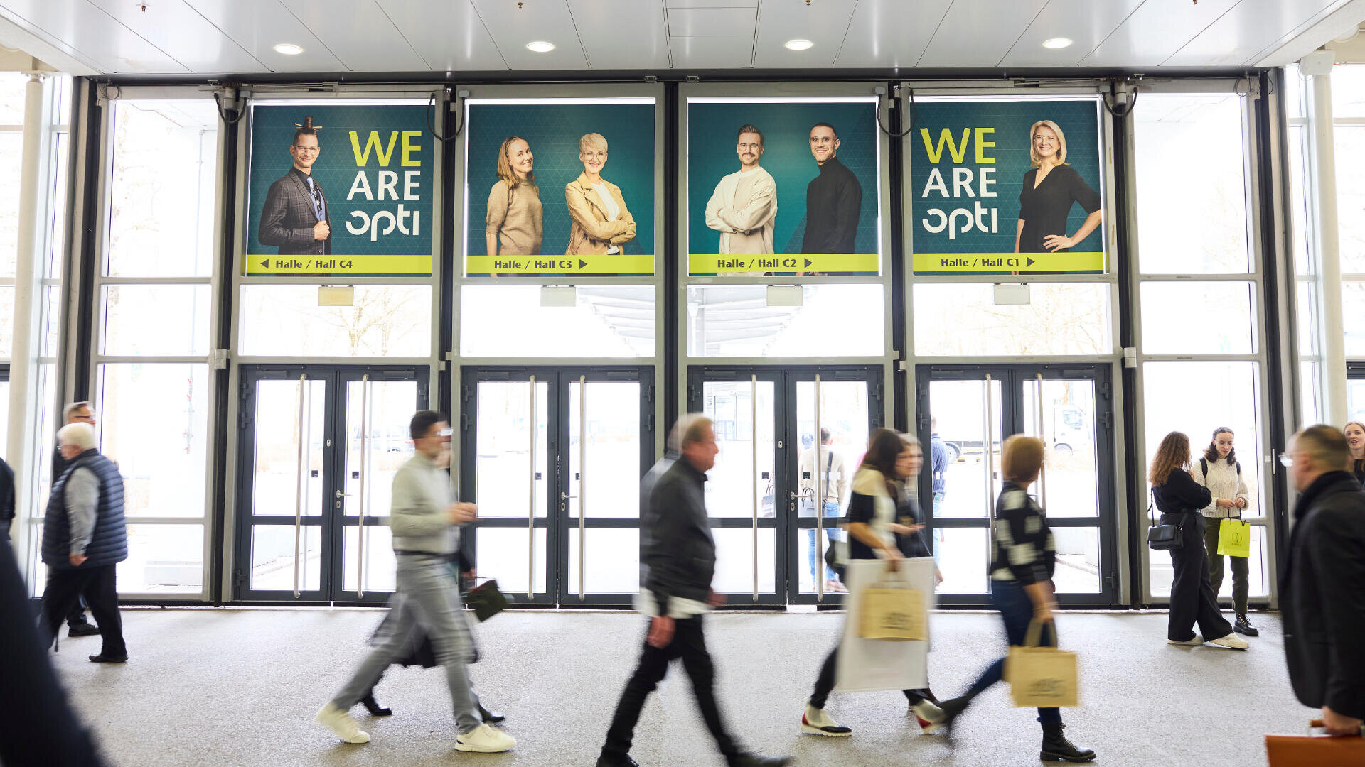 Man geht durch einen hellen, modernen Flur mit großen Glastüren. Über den Türen hängen drei Plakate mit lächelnden Menschengruppen und dem fettgedruckten Schriftzug „WIR SIND opti“.