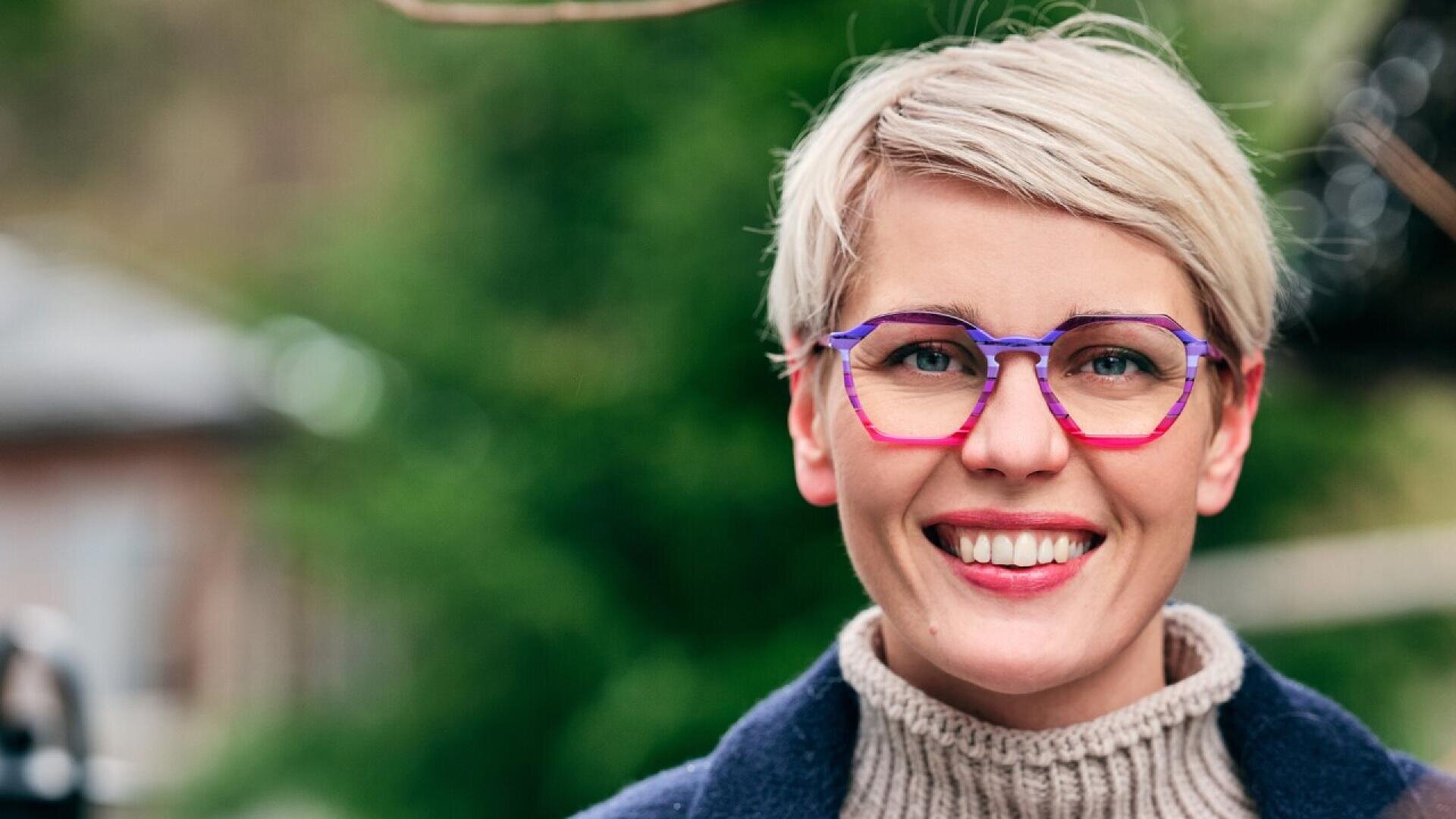 A smiling person with short blond hair and colorful glasses stands outdoors, wearing a beige turtleneck sweater and a dark coat, with a blurred green background behind them.