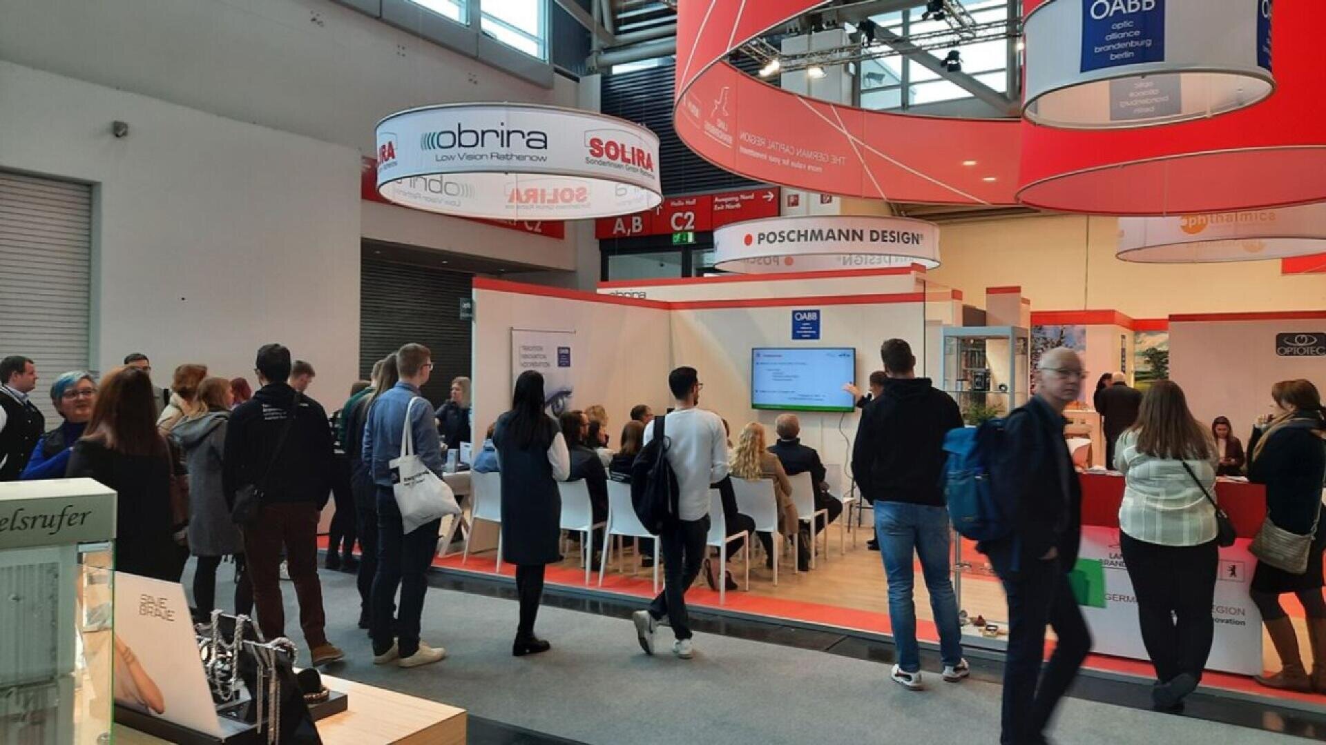 A group of people attends a presentation at a trade show or convention, seated and standing near booths with banners and display screens in a large, well-lit indoor venue.
