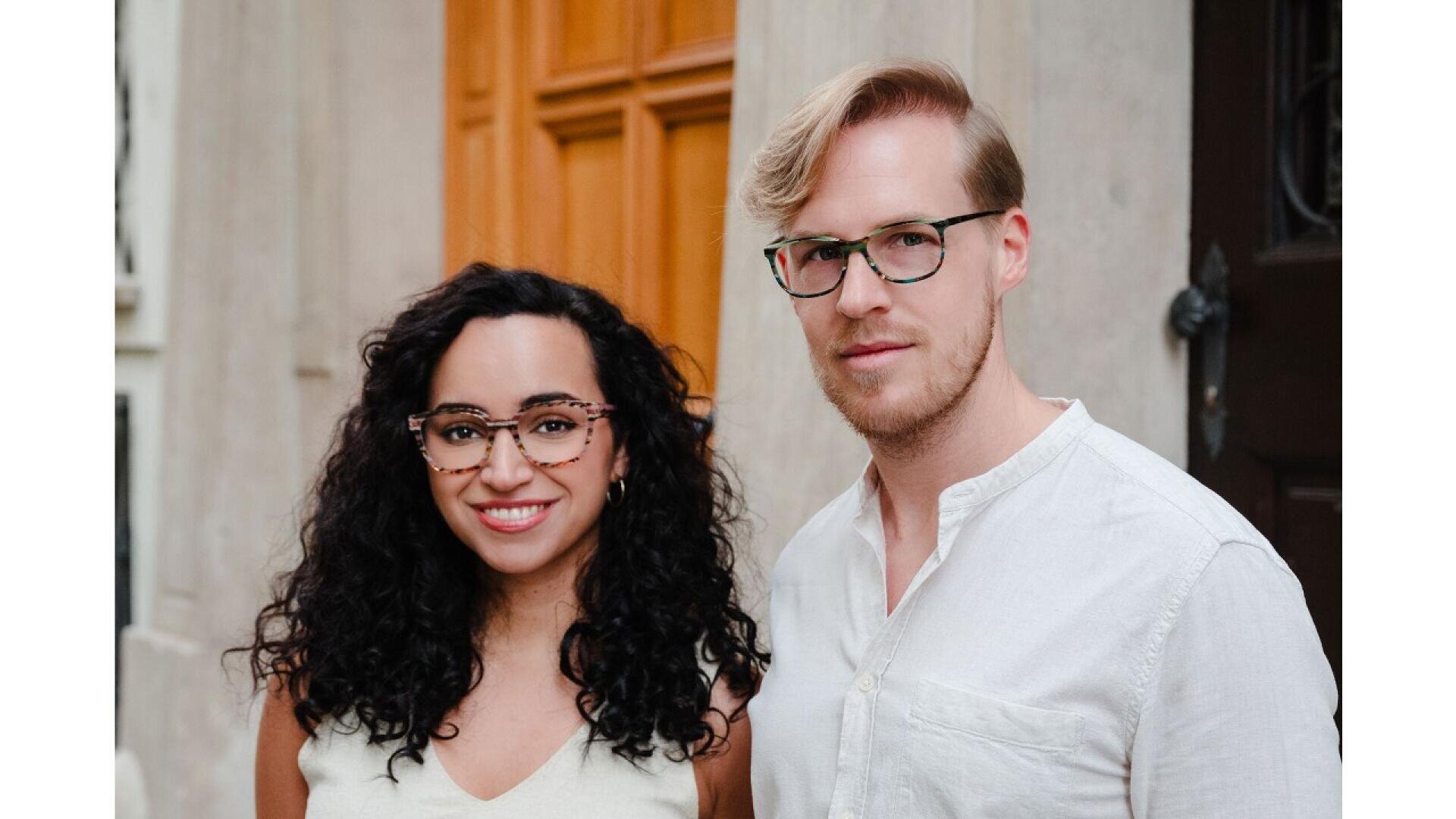 A woman with curly dark hair and a man with light hair, both wearing glasses and light-colored tops, stand side by side smiling in front of a wooden door and a stone wall.