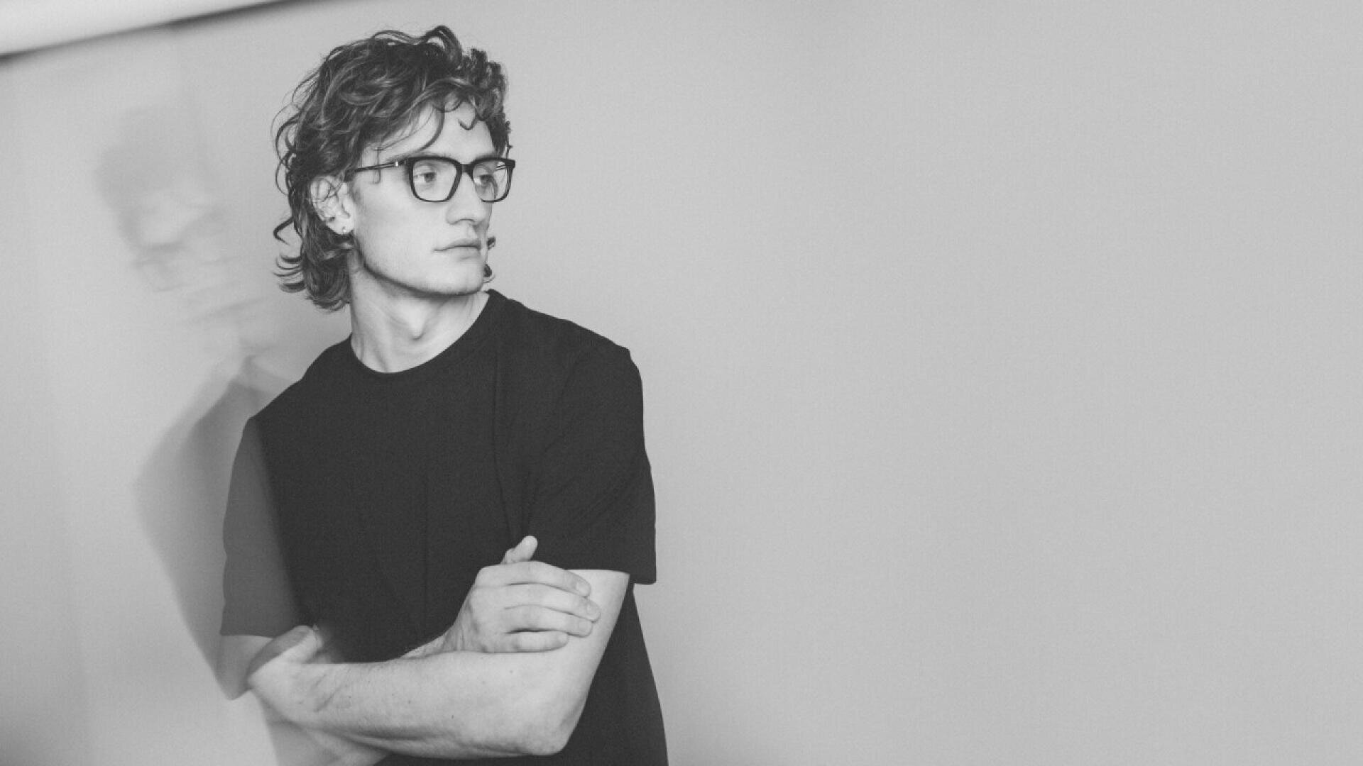 A young person with curly hair and glasses stands against a plain wall, looking to the side with arms crossed. The image is in black and white, and a faint double exposure effect shows a blurred outline of the figure.