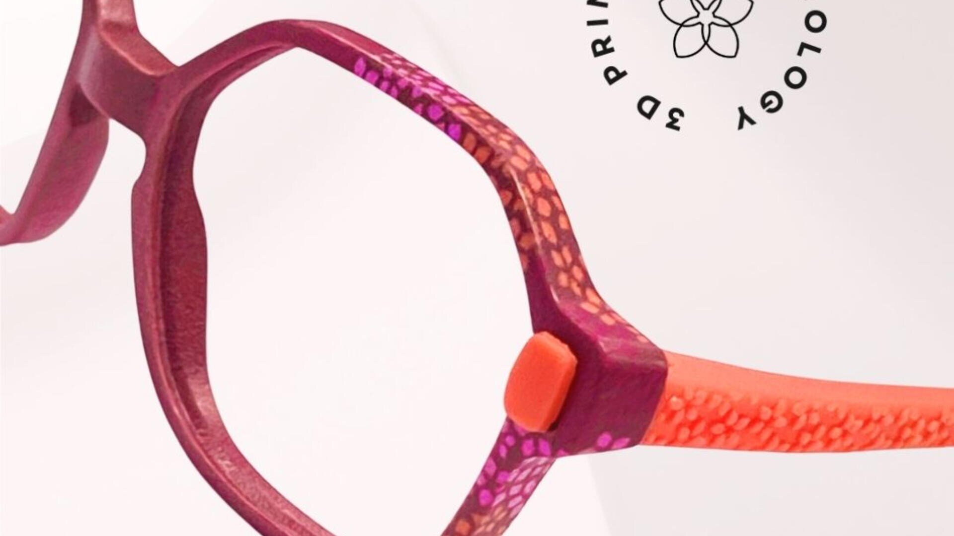 Nahaufnahme einer bunten Brillenfassung mit einer Mischung aus Lila-, Orange- und Rosatönen und einem strukturierten Muster. Im Hintergrund befindet sich ein rundes schwarzes Logo mit den Worten „3D PRINTED“ und einem floralen Design.