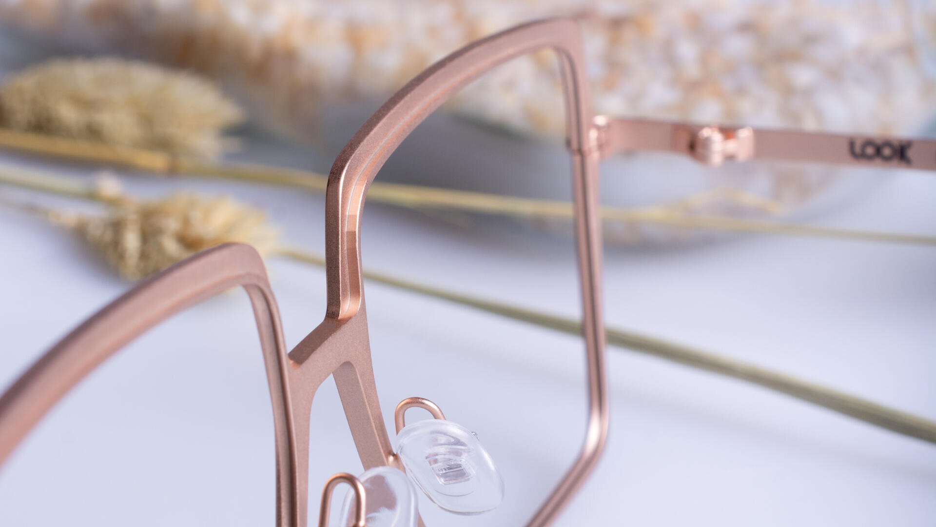 Close-up of modern rose gold eyeglasses with clear nose pads, lying on a white surface with dried wheat stems and a blurred background.