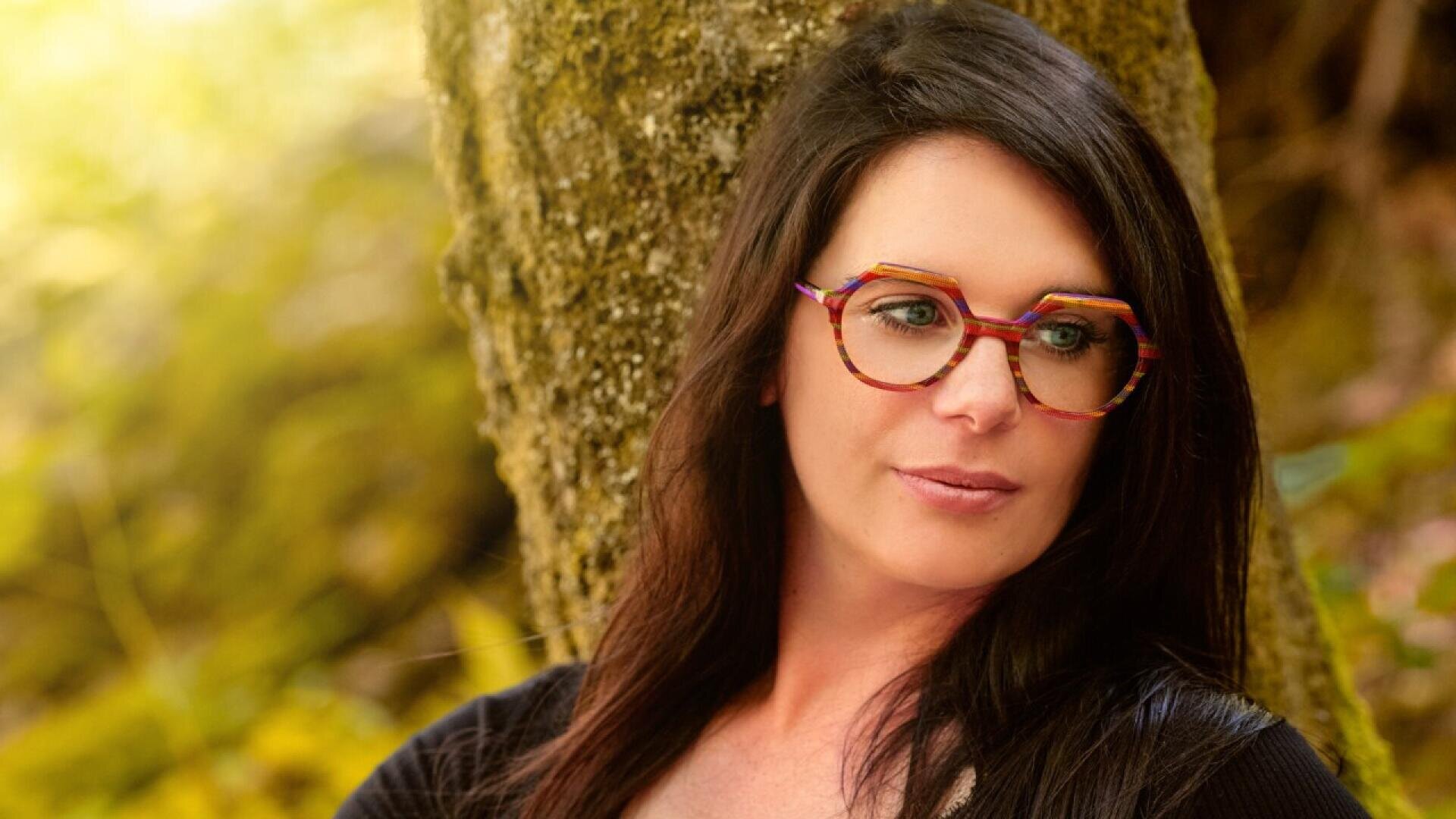 A woman with long dark hair and large, colorful glasses leans against a tree, looking to the side, with a blurred background of greenery and sunlight.