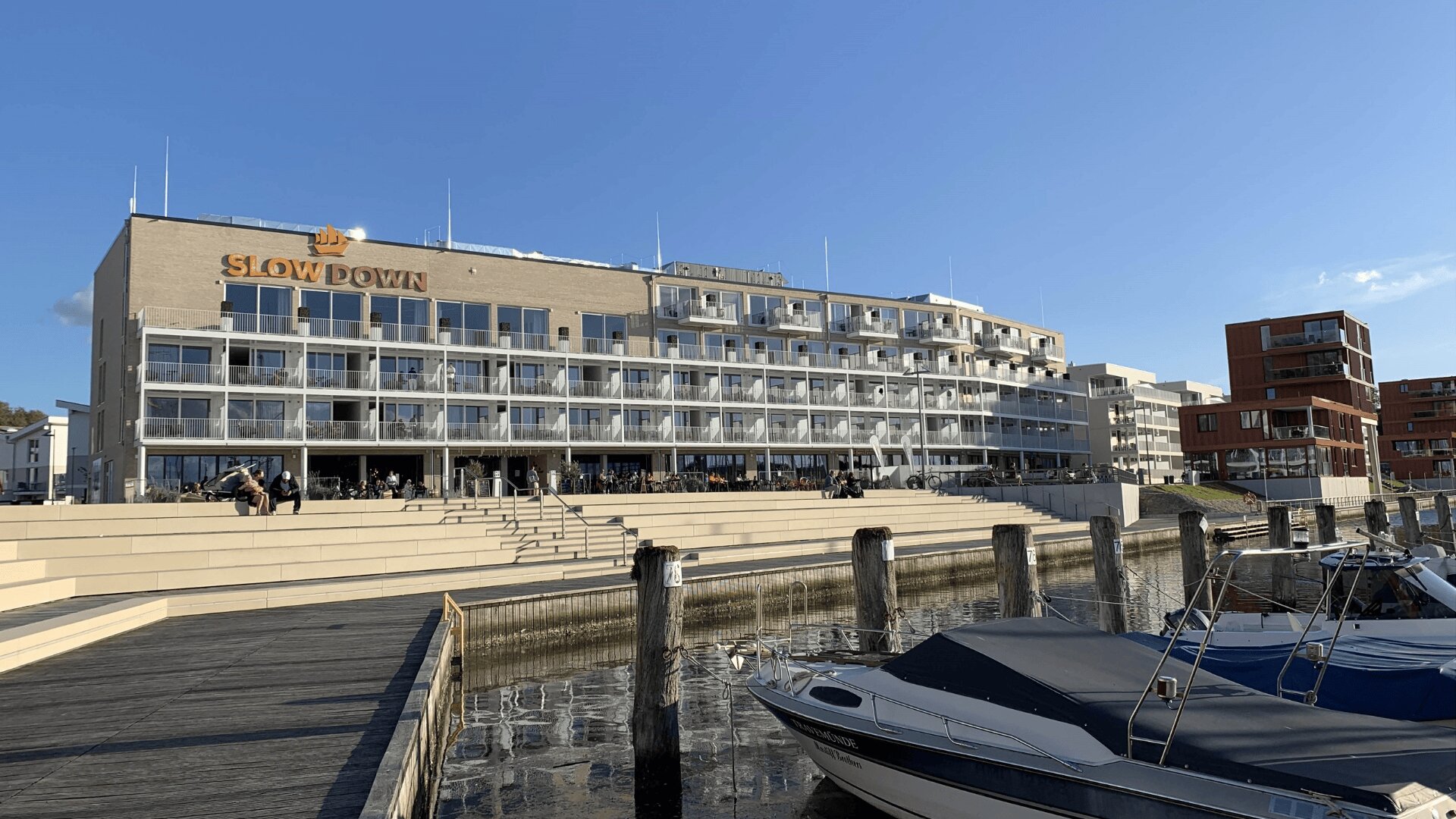 Ein modernes Hotel am Wasser namens Slow Down mit Balkonen überblickt einen Jachthafen mit angedockten Booten, Holzstegen und einer Außenterrasse unter blauem Himmel.