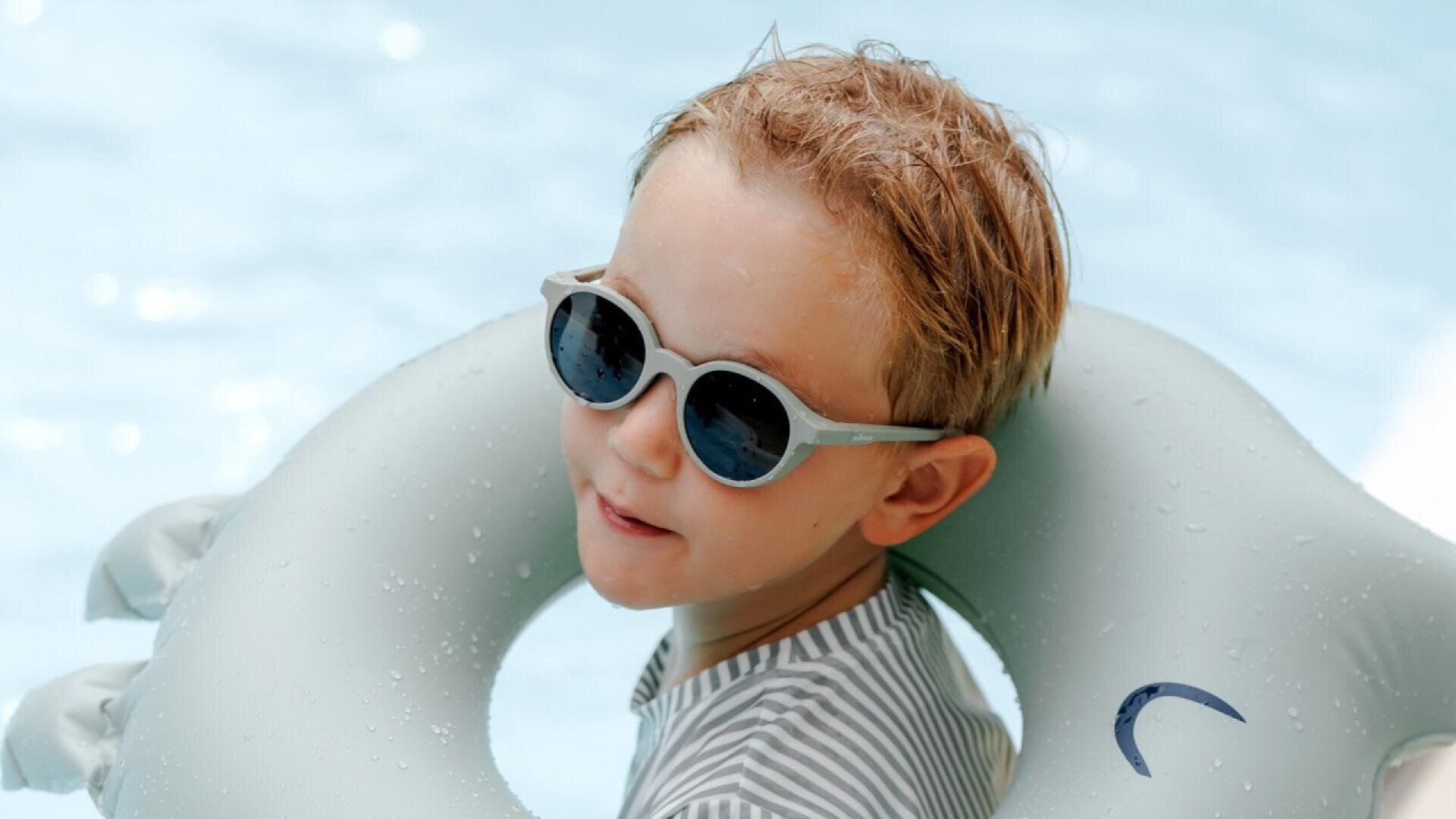 A young boy wearing striped swimwear and sunglasses smiles while floating in a pool with a light gray inflatable ring around him. The water is clear and blue in the background.